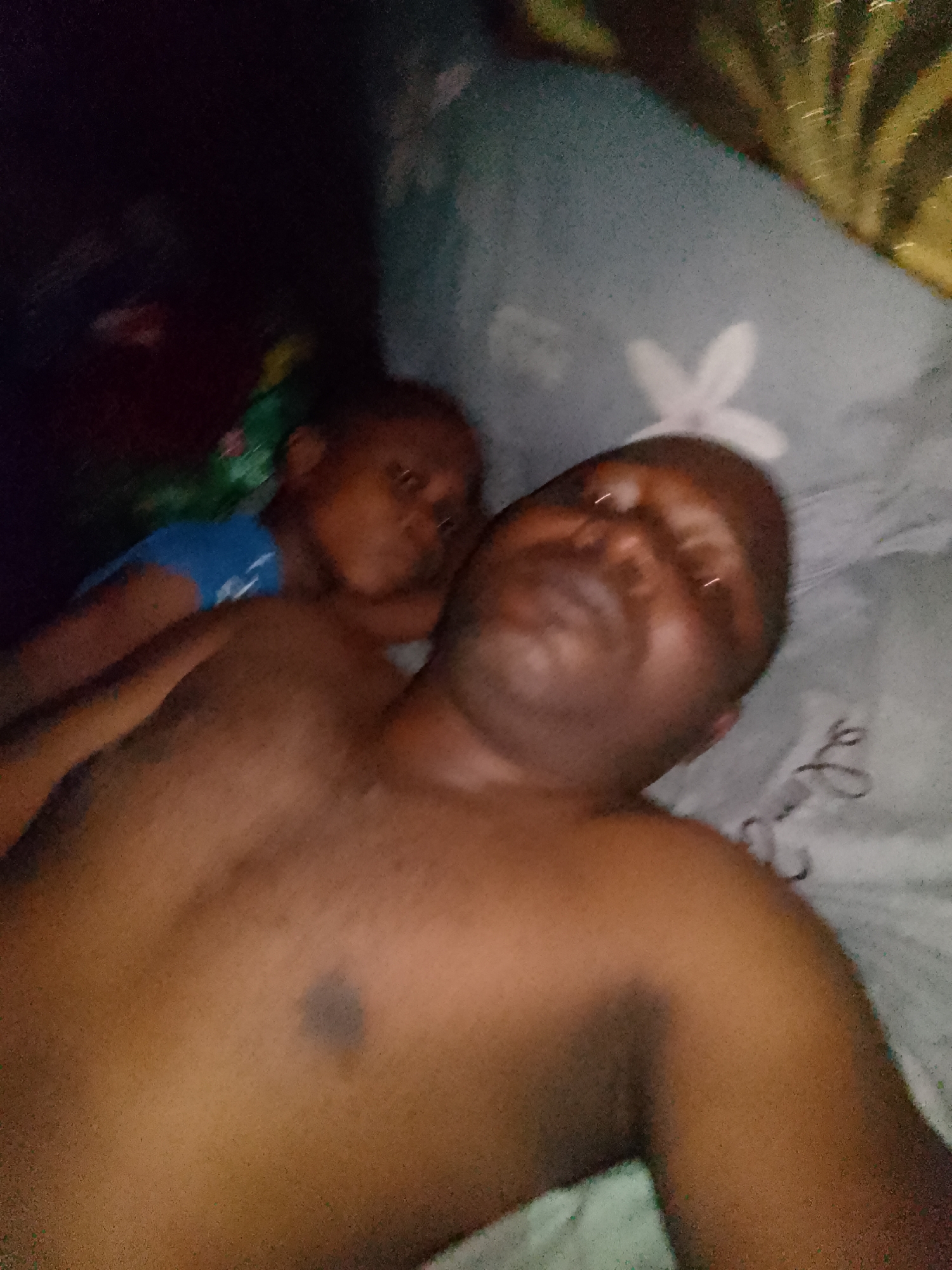 A man and a child are lying on a bed with patterned sheets. The man is shirtless and the child is wearing a blue shirt. The location is unknown.
