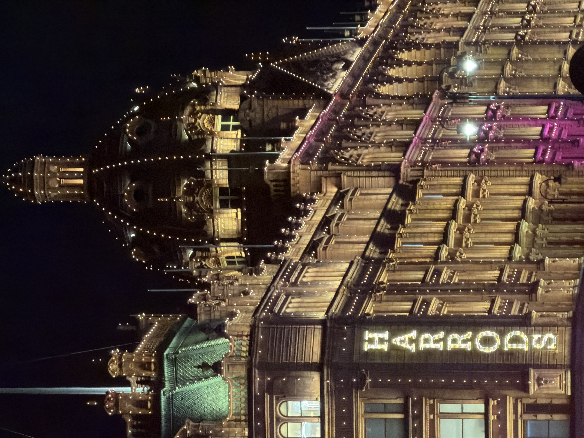 The image depicts Harrods department store in London at night. The building is elaborately decorated with lights, including strings of small lights outlining the architectural details and the store's name illuminated above the entrance. The ornate building facade is visible with the distinctive green dome, and various levels are illuminated. The sky is dark, providing a backdrop for the glowing building. There are no people visible in the image.