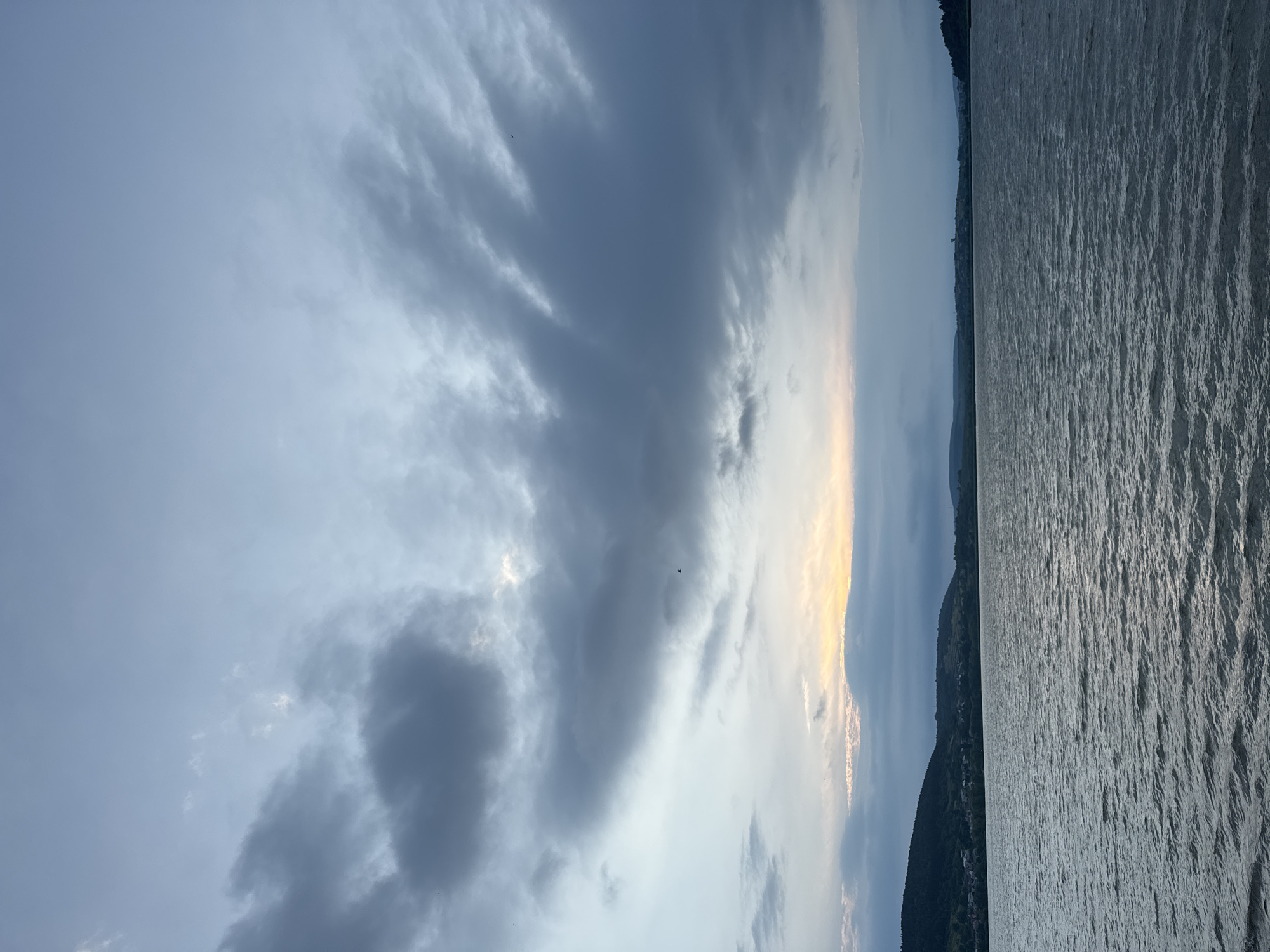 This is a vertical, outdoor, wide-angle photograph taken at dusk overlooking a large body of water, likely the Ternopil Reservoir, near Berezhany, Ukraine. The sky is filled with dramatic, dark gray clouds that are breaking in the center to reveal a pale yellow and orange glow of the setting sun. A few small birds are visible in the sky. The water in the foreground is choppy, reflecting the muted colors of the sky. In the distance, the shore is lined with dark, rolling hills covered in trees. A small settlement, with visible houses and structures, is nestled on the hillside. The overall mood is atmospheric and serene.