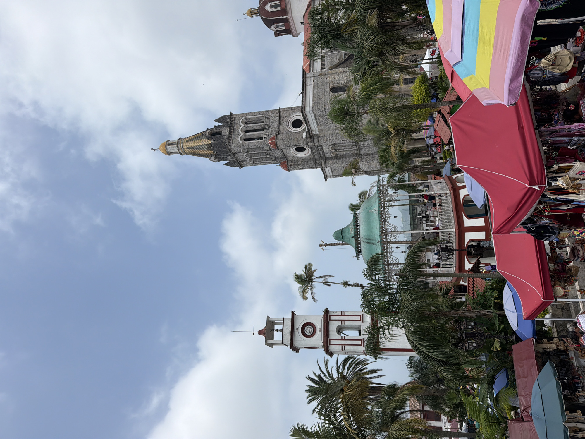 The image shows a view of Cuetzalan, Mexico, with a focus on its architectural landmarks and vibrant market scene. Three prominent towers and domes are visible, each showcasing distinct architectural styles. The first tower is white with red accents and a clock face. The second is a stone structure topped with a gold and reddish-brown conical roof. The third dome has a faded green patina, suggesting age and exposure. Lush palm trees punctuate the landscape, adding a tropical feel to the scene. In the foreground, colorful awnings from market stalls create a dynamic contrast against the historical buildings, indicating a bustling commercial activity. The sky overhead features scattered clouds, casting soft shadows that enhance the depth and texture of the scene.