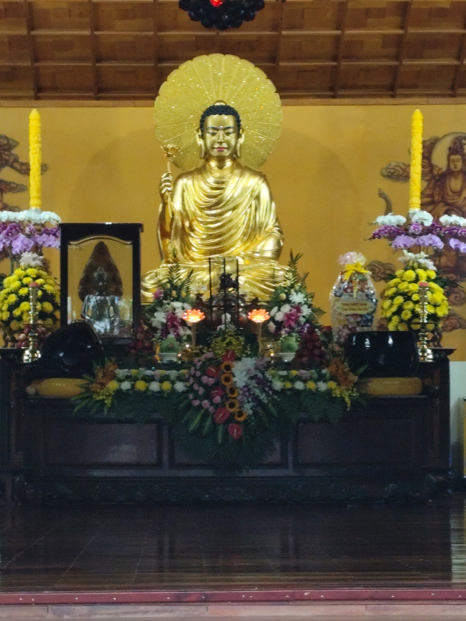 The image shows a golden Buddha statue, likely in a Buddhist temple, with offerings of flowers and other objects arranged in front of it. Two tall yellow candles flank either side of the statue. A framed image is to the left of the statue. The scene takes place in the city of Da Lat, Vietnam. The statue is seated, with a golden halo-like disc behind the head. There is a wooden alter and the floor is made of wood. There are decorations of flowers and fruit on the alter.