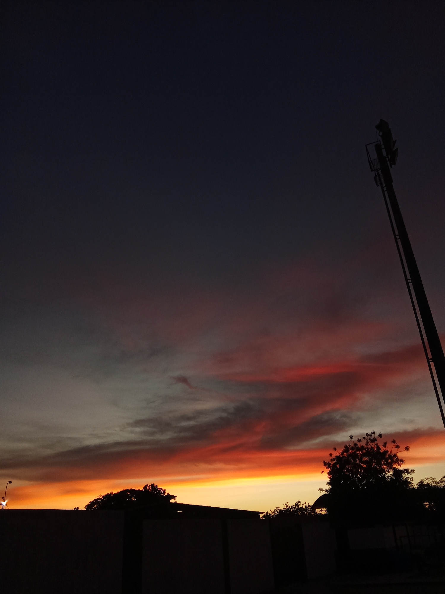 A dusk scene in Juan Griego, Venezuela presents a striking contrast between a fiery orange and red sunset horizon and a darkening blue sky. The silhouette of a tree and a utility pole with mounted equipment are visible against the twilight sky. A low wall runs across the bottom portion of the frame. A lone street lamp illuminates the lower left corner. The overall atmosphere is one of fading light and encroaching darkness.