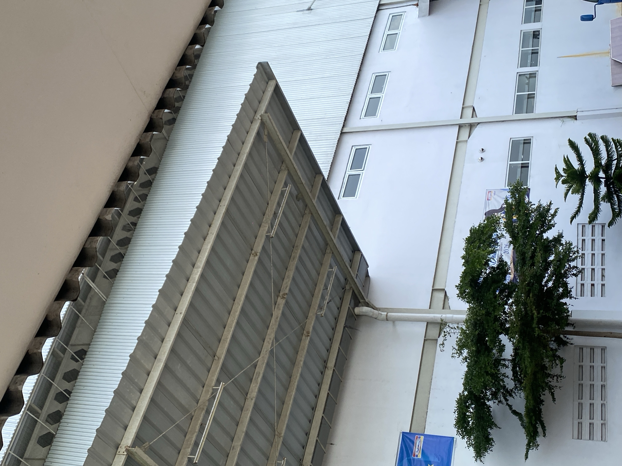 The image depicts the exterior of a white building, likely an industrial facility, in Gambut Barat, Indonesia. A corrugated metal overhang supported by beams extends from the building, casting a shadow on the wall below. A row of evenly spaced rectangular windows is visible on the building's facade. In the foreground, a lush green tree with dark foliage is situated to the left, partially obscuring some of the windows and a white rectangular vent on the wall. To the right of the tree, a smaller, spiky evergreen tree is visible. A blue banner with text and a logo is partially visible on the wall to the left of the tree. The sky is overcast and appears bright. The architectural style suggests a modern industrial or commercial structure.