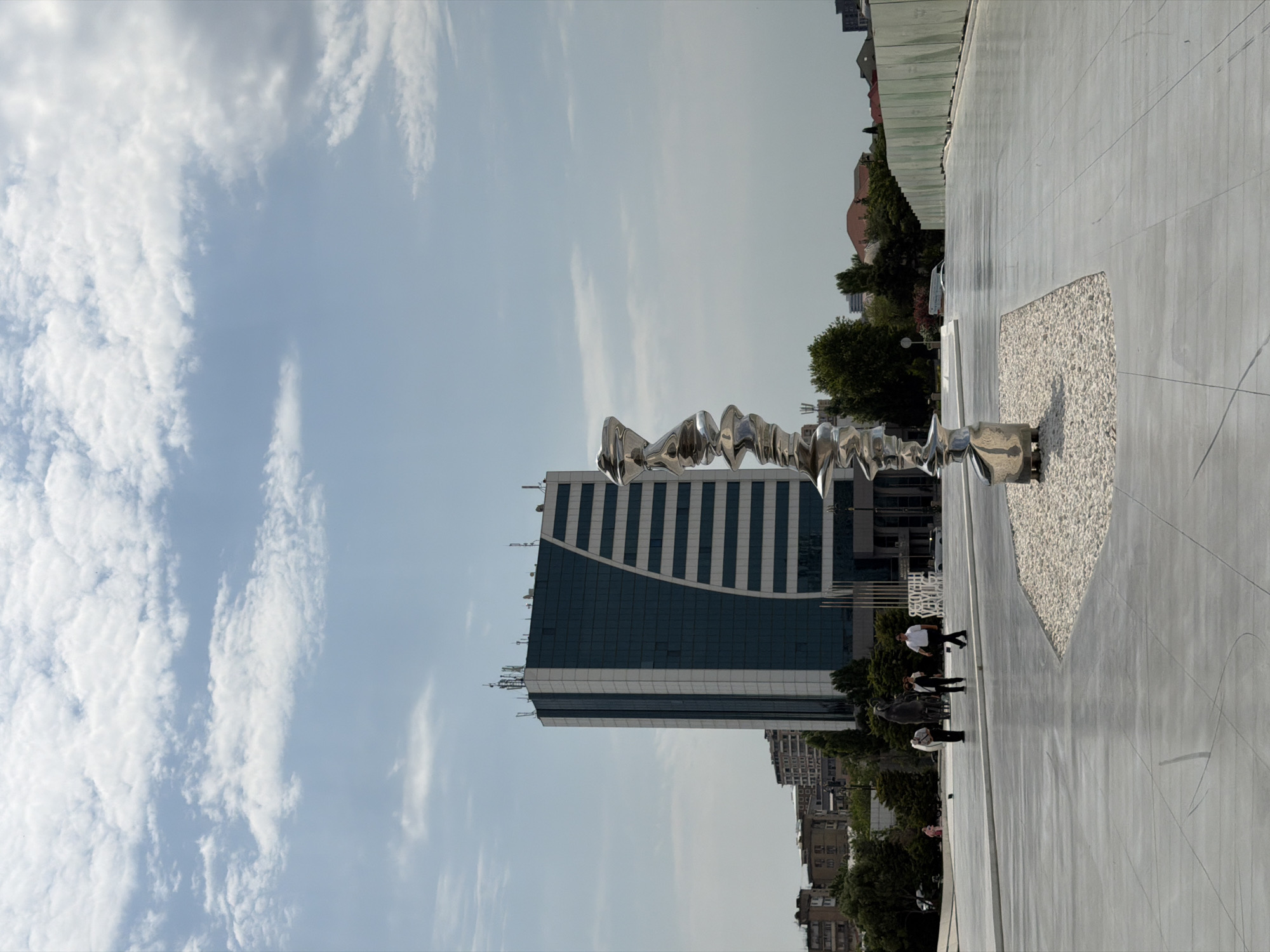 The image captures a bright, daytime scene in Baku, Azerbaijan, likely at the Baku Congress Center, given the architectural style and the presence of contemporary art. In the foreground, a highly polished, abstract metal sculpture with a spiral form stands prominently on a bed of small white pebbles. The sculpture reflects the surrounding environment and the sky. To the left of the sculpture, a wide, glossy grey paved plaza extends outwards, with some of the paving tiles showing subtle parallel lines.
In the mid-ground, several individuals are seen walking across the plaza. Two men in the center are walking away from the viewer, one in a dark suit and the other in a lighter colored jacket and pants. To their left, a man in a white shirt and dark pants walks towards the viewer, while another person in a dark outfit is further left. In the background, a modern, tall building with a dark glass facade and white horizontal banding dominates the skyline. A curved section of white and green horizontal bands is visible on the right side of this building. To the left of this main building, older, multi-story residential buildings with a more traditional architectural style are visible, along with lush green trees. The sky above is a light blue with scattered white cumulus clouds. A long, light green fence runs along the right edge of the mid-ground, separating the plaza from an unseen area.