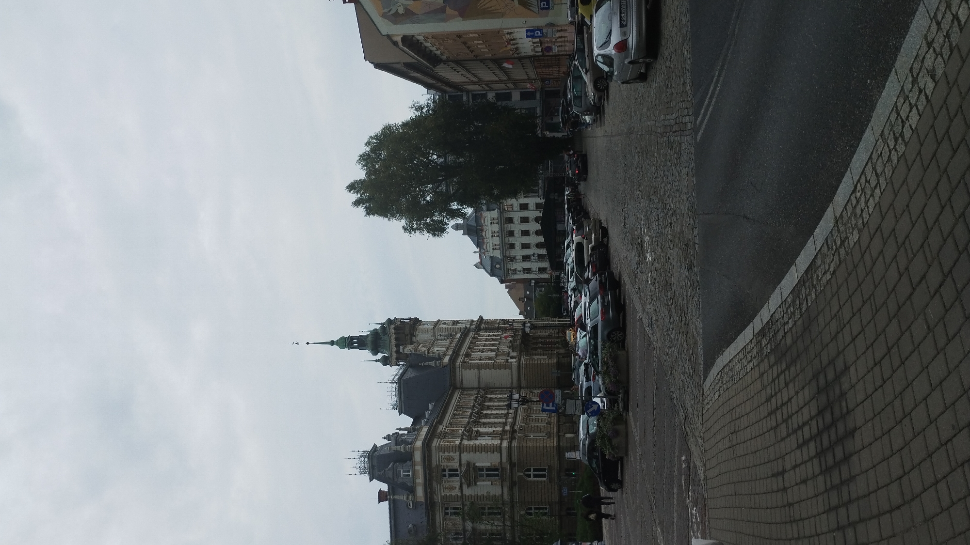 The image depicts a street scene in Bielsko-Biala, Poland, likely in a historic city center. In the foreground, a cobblestone sidewalk leads to a paved road. On the left side of the road, a large, ornate building with a distinctive tower and mansard roof dominates the view. This building appears to be of historical significance, possibly a government building or a grand residence. Several cars are parked in a lot in front of this building and along the street. Across the street, another building with a more modern facade, featuring many windows, is visible. Further down the street, a tall, leafy tree stands out against the overcast sky, and more buildings line the right side of the road, including one with a mural on its side. Several parked cars are visible throughout the scene, and a few pedestrians are walking on the sidewalk. The overall atmosphere is that of a typical European city square on a cloudy day.