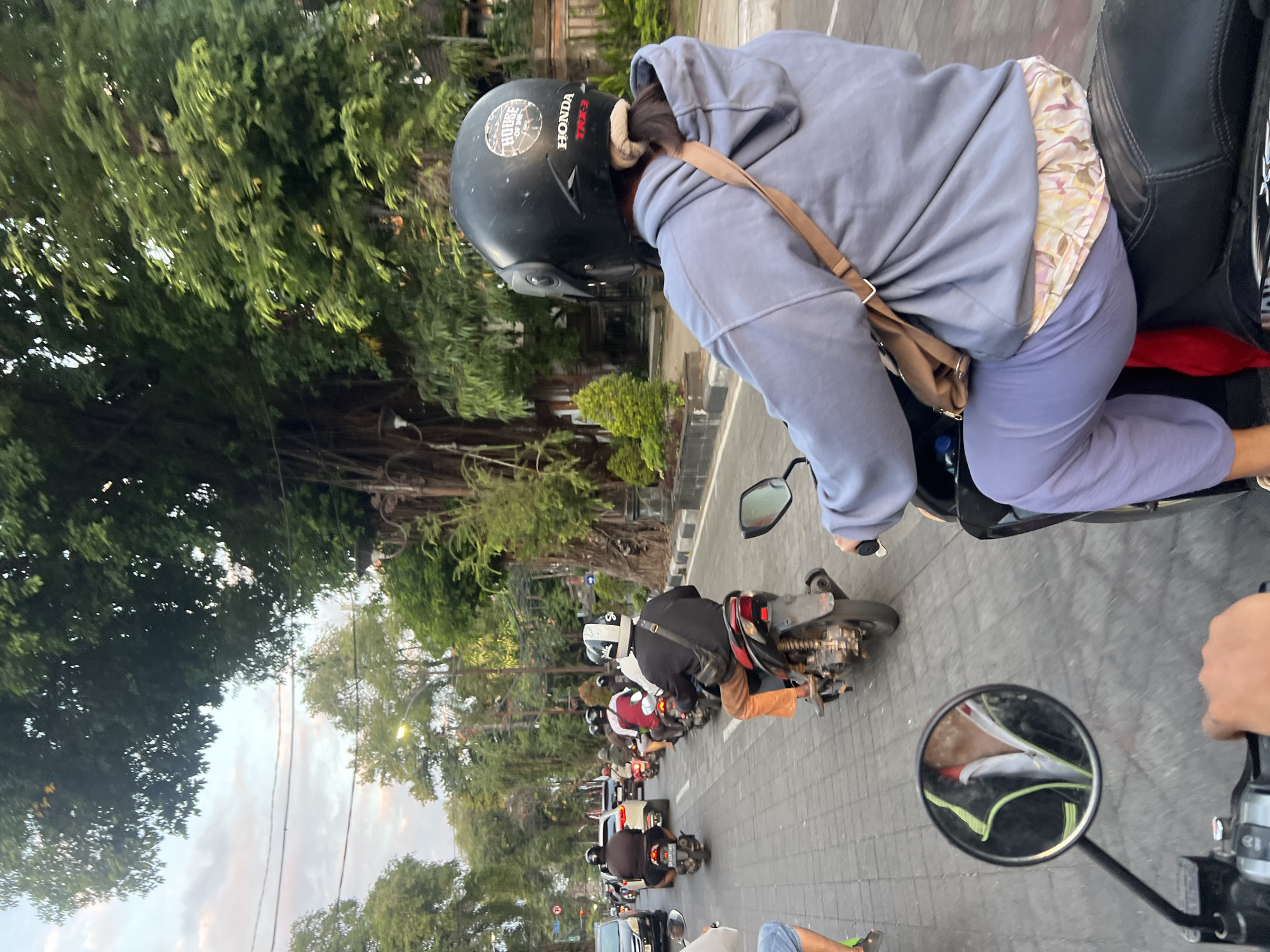 The image depicts a congested street scene in Denpasar, Indonesia, likely during twilight hours. The foreground is dominated by the back of a person riding a scooter, wearing a grey hoodie and light purple pants, with a brown crossbody bag. The scooter has a black seat and chrome accents. A helmet, black with white and red accents, is on the rider's head, displaying a "HOUSE OF DJ" sticker and "HONDA" branding.
In the mid-ground, a line of scooters and motorcycles is moving along a paved road. One rider in front is wearing orange pants and a black jacket. Other vehicles are visible in the background, including cars. The street is lined with trees, some with large, gnarled trunks, and there are also decorative iron fences and some low-lying greenery. The sky above is a pale blue with hints of orange and pink, suggesting either dawn or dusk. The overall atmosphere is one of busy urban traffic. The presence of "Honda" branding on the scooter and the general ambiance suggest a typical street scene in a Southeast Asian city.