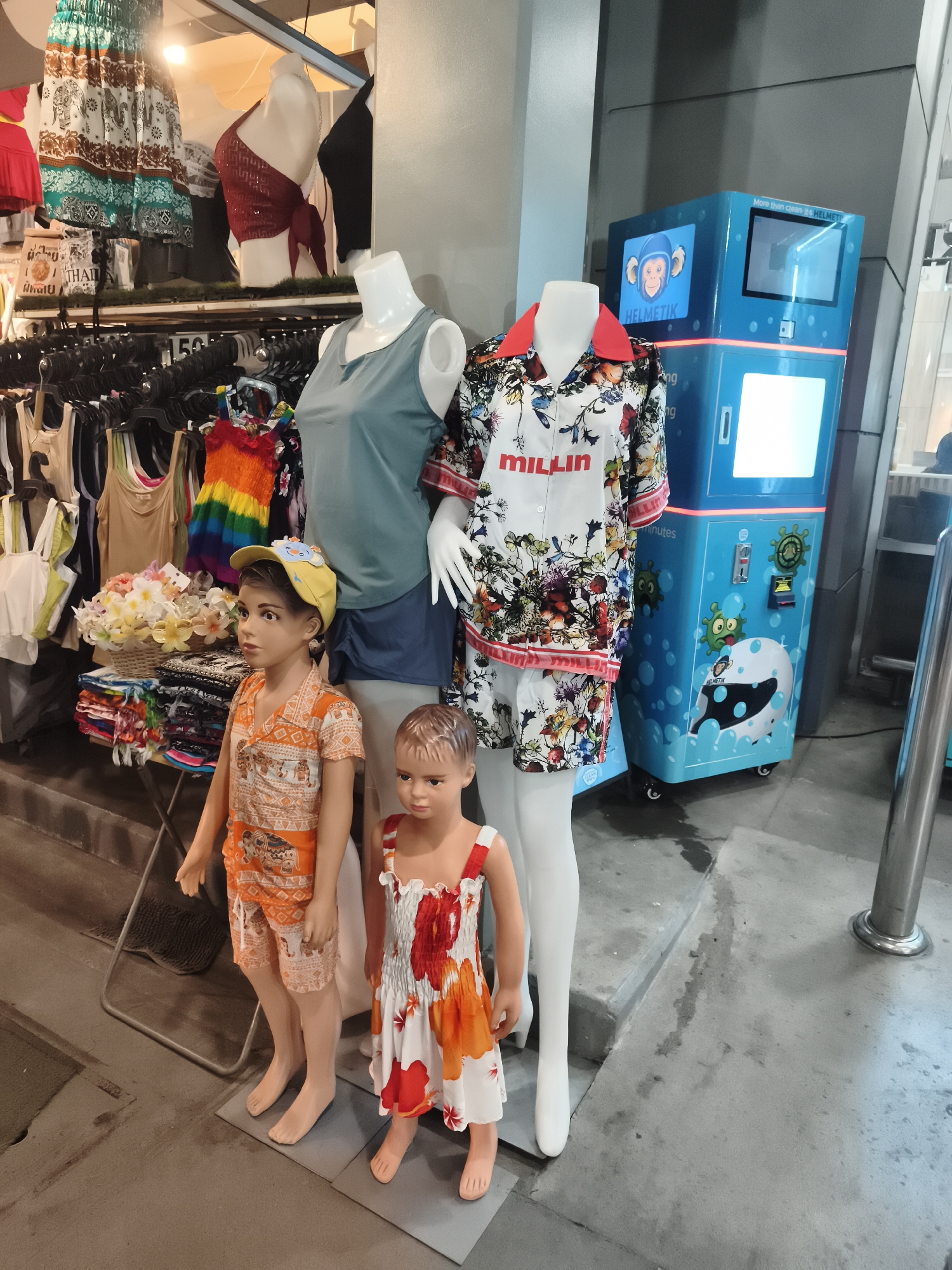 The scene captures a clothing store display in Baan Lamai, Thailand. Two mannequins, one with a light blue sleeveless top and blue shorts, and the other wearing a white shirt and short set with a floral pattern and red trim, are positioned next to child mannequins. The child mannequin on the left wears an orange outfit with elephant patterns and a yellow cap, while the other wears a white dress with orange and red floral accents. Behind the mannequins, clothing items hang on racks, with skirts and tops visible. To the right is a helmet cleaning machine with a blue exterior, featuring cartoonish images and the text "HELMETIK" and "More than clean, it's HELMETIK." A metallic pole is visible on the far right. The setting suggests a shopping area, likely frequented by tourists.