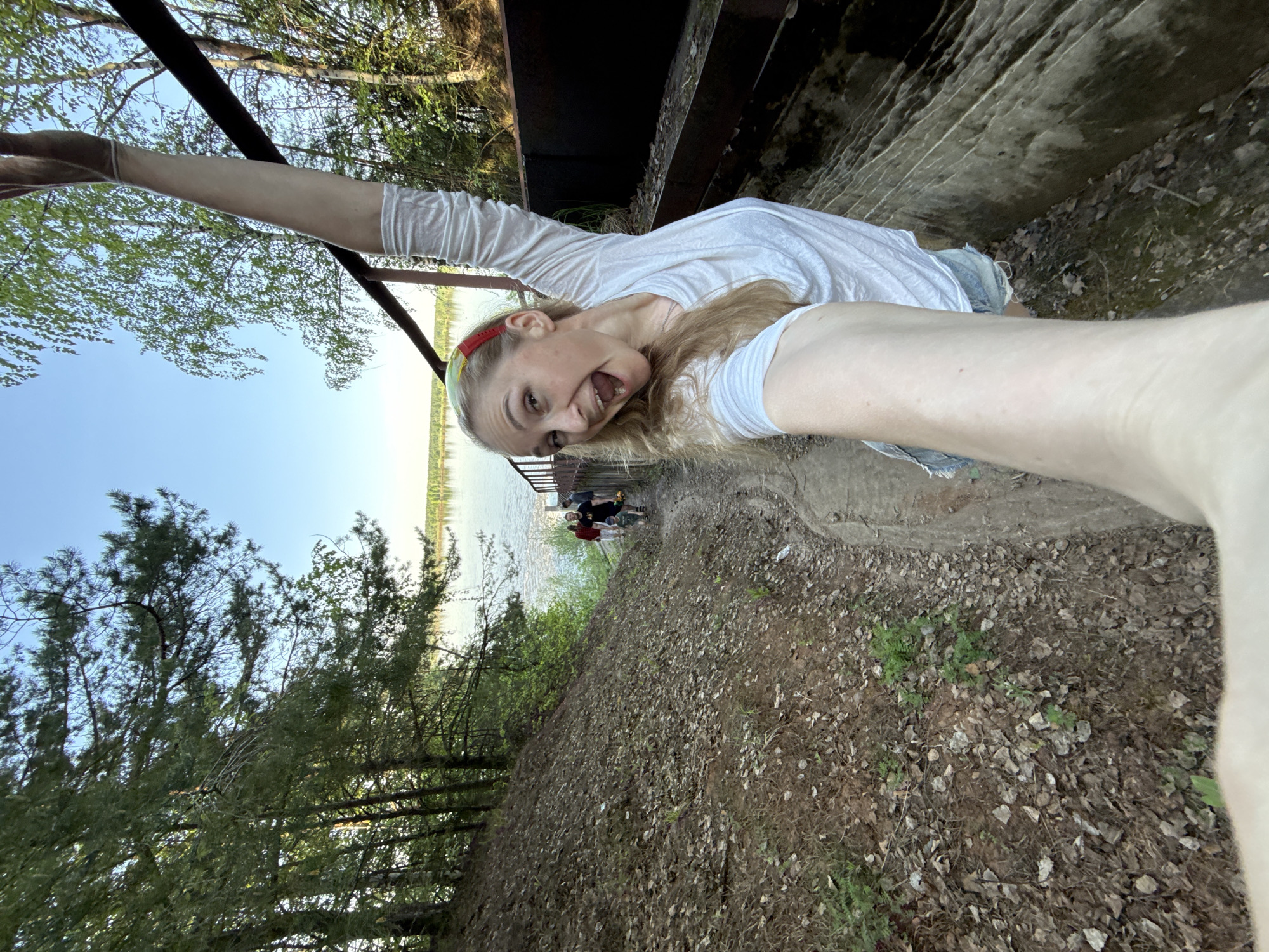 A woman takes a selfie outdoors, appearing to hang from a structure near water. She has blonde hair, a red headband, and wears a white shirt and denim shorts. She is looking at the camera with an exaggerated expression. The scene includes trees, a body of water, and a path where other people are visible in the background. The exact location in the city of Novyi, Russia is not discernible.