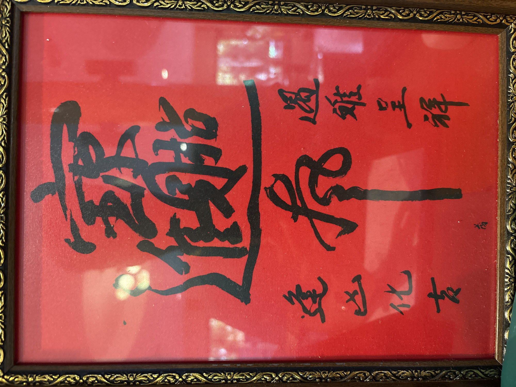 This is a close-up, eye-level shot of a framed piece of calligraphy on a vibrant red background. The calligraphy is done in bold black ink, appearing to be traditional Chinese or Japanese characters. The frame itself is ornate, with a dark wood base and intricate gold detailing along its edges. The calligraphy is arranged in several vertical lines. The dominant, largest characters appear in the upper half of the frame, followed by smaller characters arranged in columns to the left and right of a central, thick vertical stroke. The overall impression is one of cultural significance and decorative artistry. The specific location in Chalong, Thailand, is not discernible from the image itself, which focuses solely on the framed artwork.