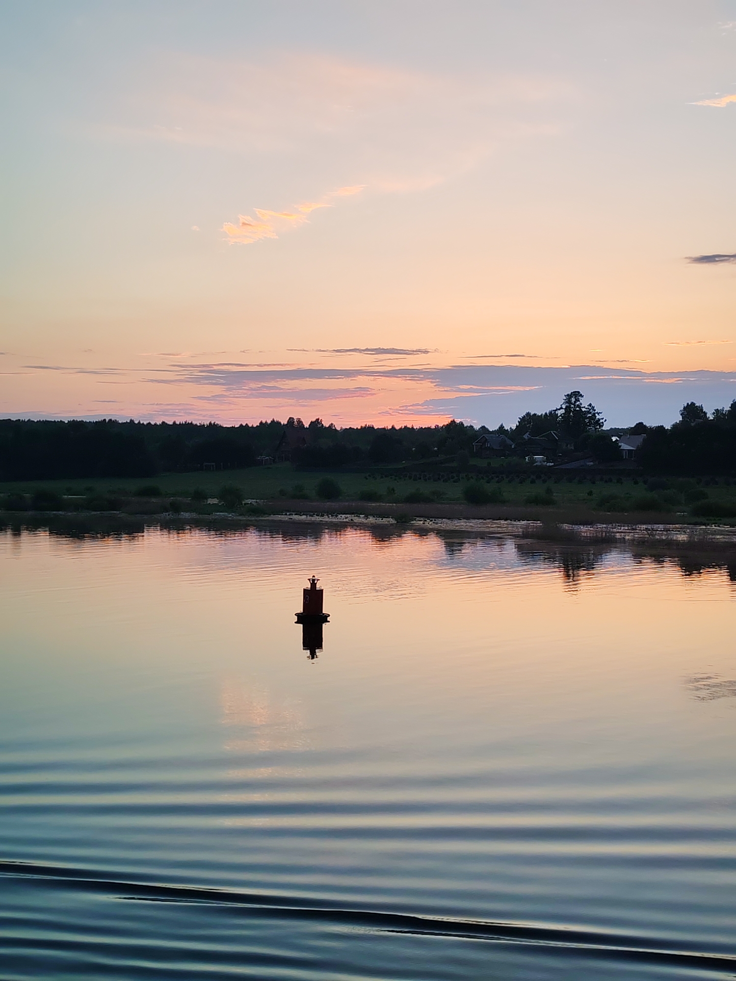 The image depicts a tranquil body of water at sunset in Unknown, Unknown. A red navigation buoy is centrally located in the water, with its reflection visible on the surface. The water displays gentle ripples, creating horizontal lines. The shoreline is lined with trees and foliage. In the background, several houses or buildings are visible. The sky is a blend of light blue, pink, and orange hues, indicating the sunset.
