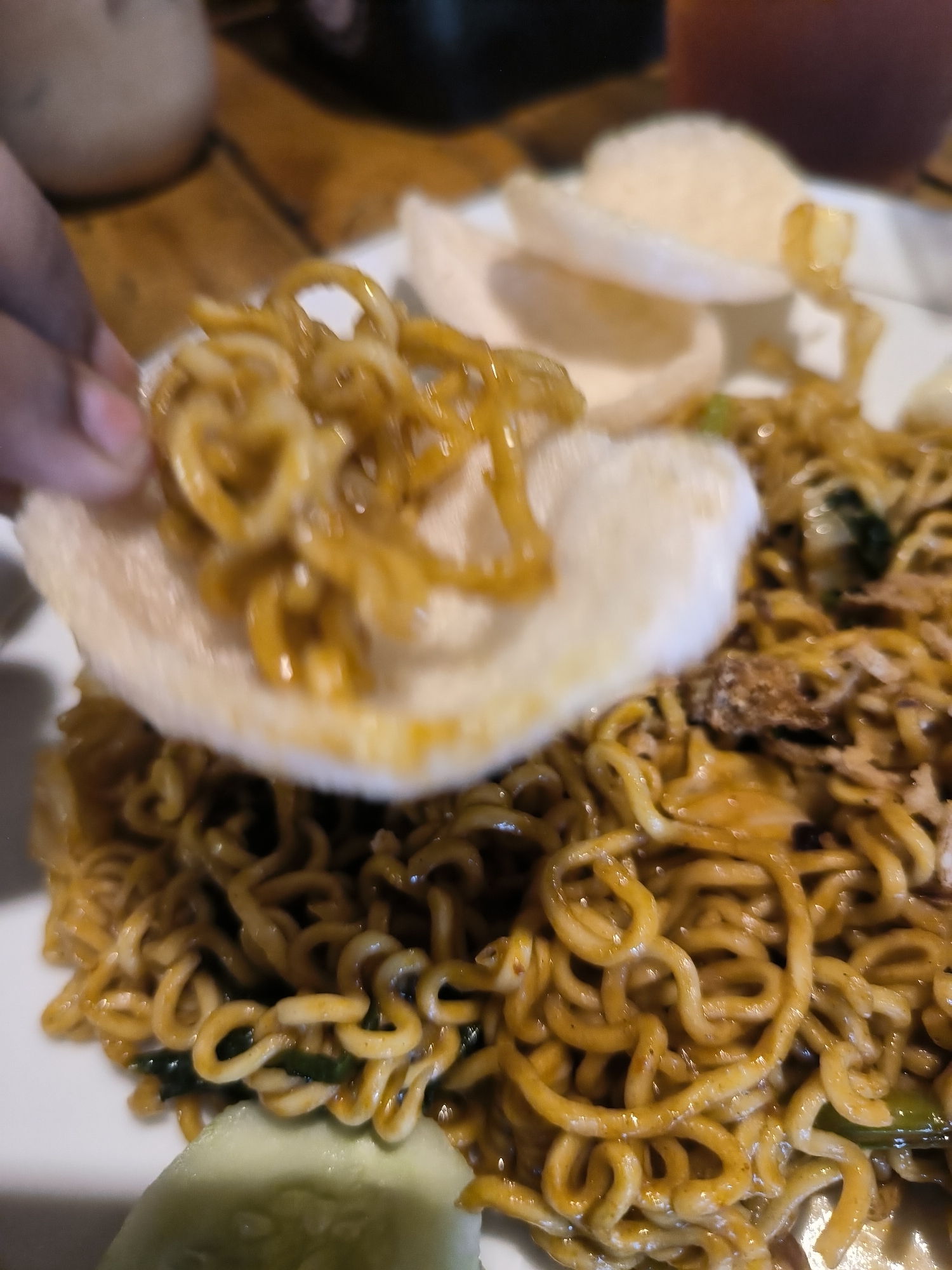 A close-up shot shows a plate of mie goreng (fried noodles) with prawn crackers. One cracker is being held up, with noodles placed on top. Cucumber slices are visible in the lower left corner of the plate. The food appears to be served at a restaurant or cafe setting.