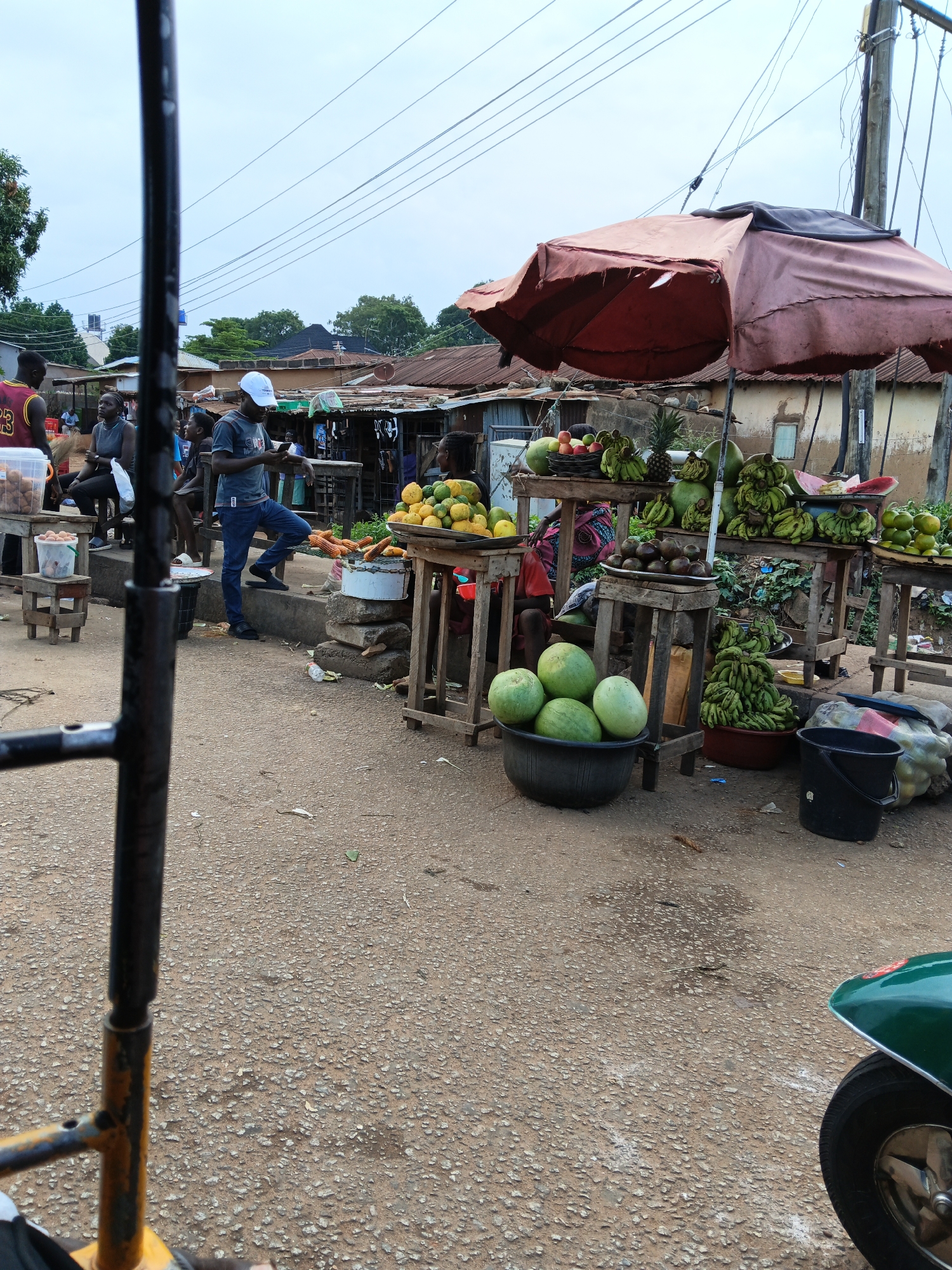 The image captures a street scene in Bukuru, Nigeria, featuring a vibrant fruit and vegetable market. In the foreground, a portion of a vehicle, possibly a tricycle, is visible, suggesting the photographer is within or near one. The market is set up along a dirt road, with numerous stalls displaying an abundance of fresh produce.
Several people are present. On the left, a man in a maroon and yellow jersey stands, while a woman in a dark grey top sits nearby. To their right, a man wearing a white cap and a blue t-shirt is looking at his phone. Other individuals are partially visible behind stalls, suggesting a bustling commercial environment.
The stalls are laden with a colorful array of fruits, including watermelons, pineapples, bananas, and what appear to be mangoes and other citrus fruits. Some vegetables, like corn, are also displayed. The produce is arranged on wooden tables and in baskets, under a large, weathered brown umbrella that provides shade.
The background shows a collection of buildings with corrugated iron roofs, typical of many urban areas in Nigeria. Overhead, a network of electrical wires crisscrosses the sky, a common sight in the region. The overall atmosphere is one of daily commerce and community life in Bukuru.