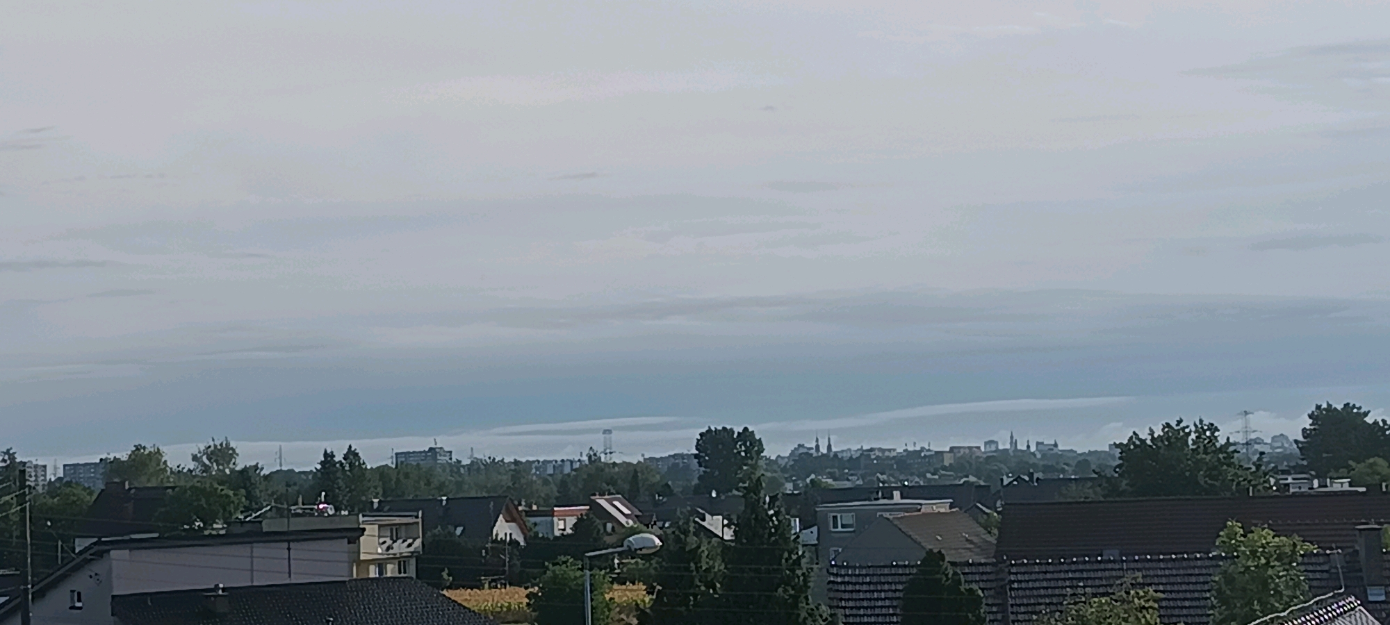 The image depicts a hazy, overcast sky over a cityscape that appears to be Opole, Poland. In the foreground, several residential buildings with pitched roofs are visible, some featuring dormer windows. Trees and foliage are interspersed throughout the residential area. In the middle ground, a denser collection of buildings and trees forms the urban landscape. The background features a more distant city skyline with taller structures and what appear to be communication towers or power lines. The overall atmosphere is muted due to the overcast conditions.
