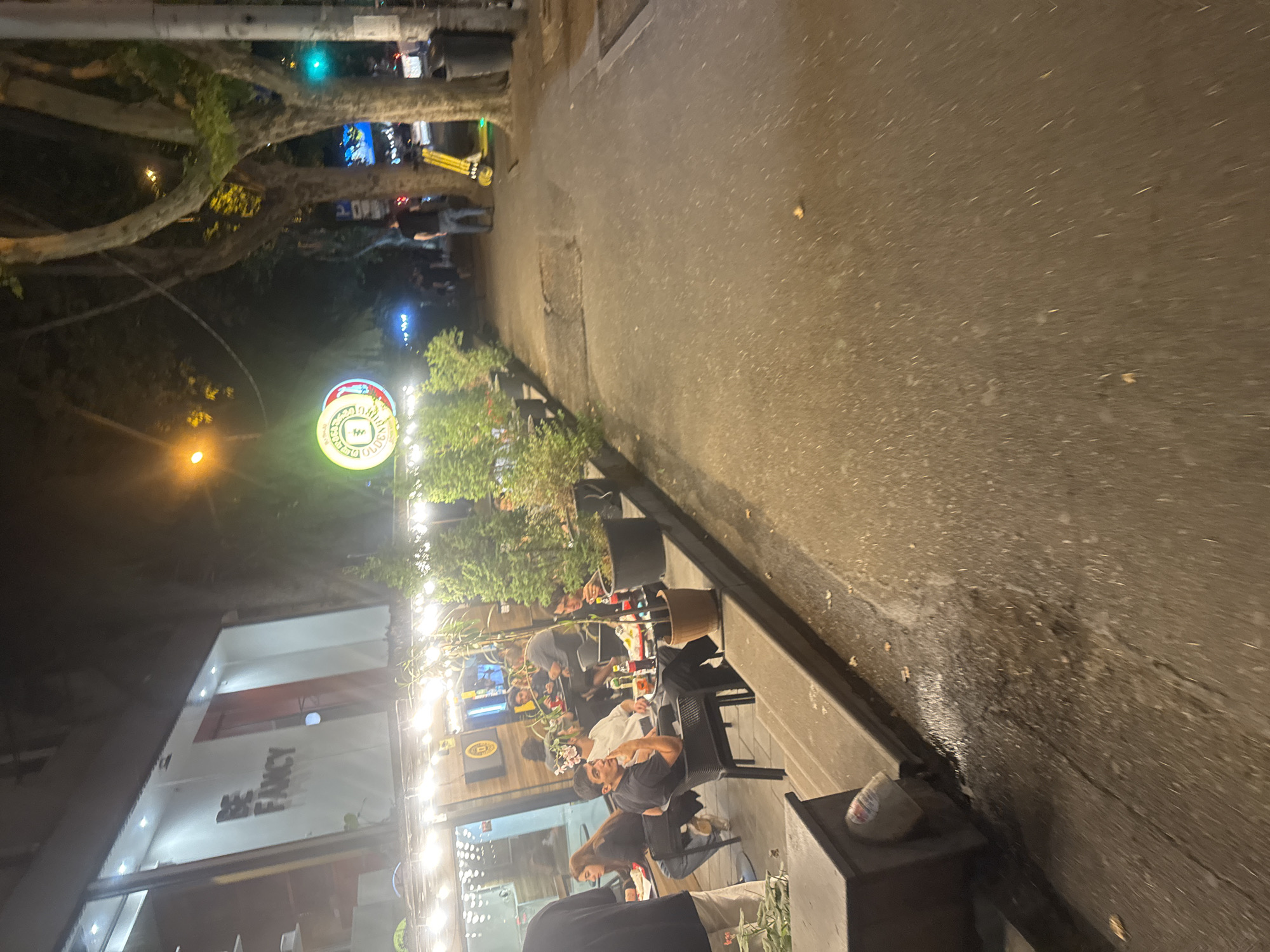 The image shows a street scene at night in Tbilisi, Georgia, likely in an area with restaurants and businesses. On the left, there is a building with a sign that reads "BE FANCY". In front of the building, an outdoor seating area for a restaurant is set up with several people dining at tables. String lights are illuminating the area. To the right of the restaurant, a sidewalk runs alongside a street lined with trees. A yellow electric scooter is parked on the sidewalk, and a person is walking away from the camera down the sidewalk. In the background, other businesses and streetlights are visible. The overall impression is a lively urban evening scene.