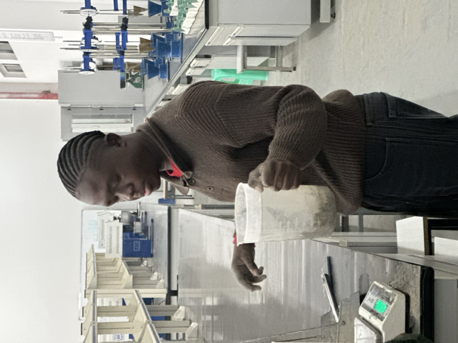 In a brightly lit laboratory, a man with braided hair is focused on pouring a substance from a large, translucent plastic container. He wears a brown button-down sweater over a red shirt and dark jeans. The lab features rows of equipment, including a weighing scale with a green display, funnels, and various industrial devices. The man's posture and gaze are directed towards the task at hand, suggesting precision and attentiveness.