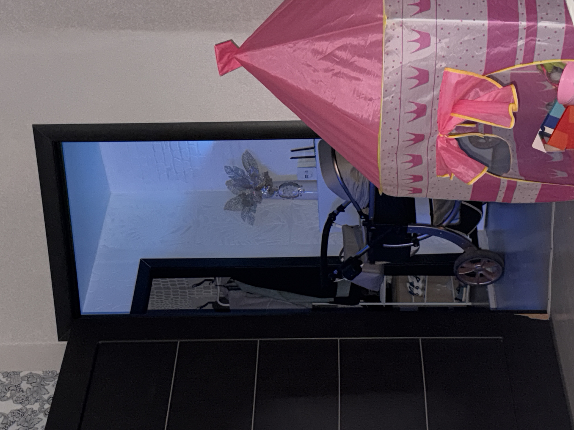 The image shows a black framed window looking into a room. Within the room, there is a baby stroller parked indoors. The stroller is located in front of a light blue wall with some decorations or objects on it. There is a pink princess-themed play tent set up next to the stroller. The picture is taken from within an adjacent room, with a partially visible patterned wallpaper on the left side and a dark paneled door or cabinet below the window. The location is Noviy, Russia.