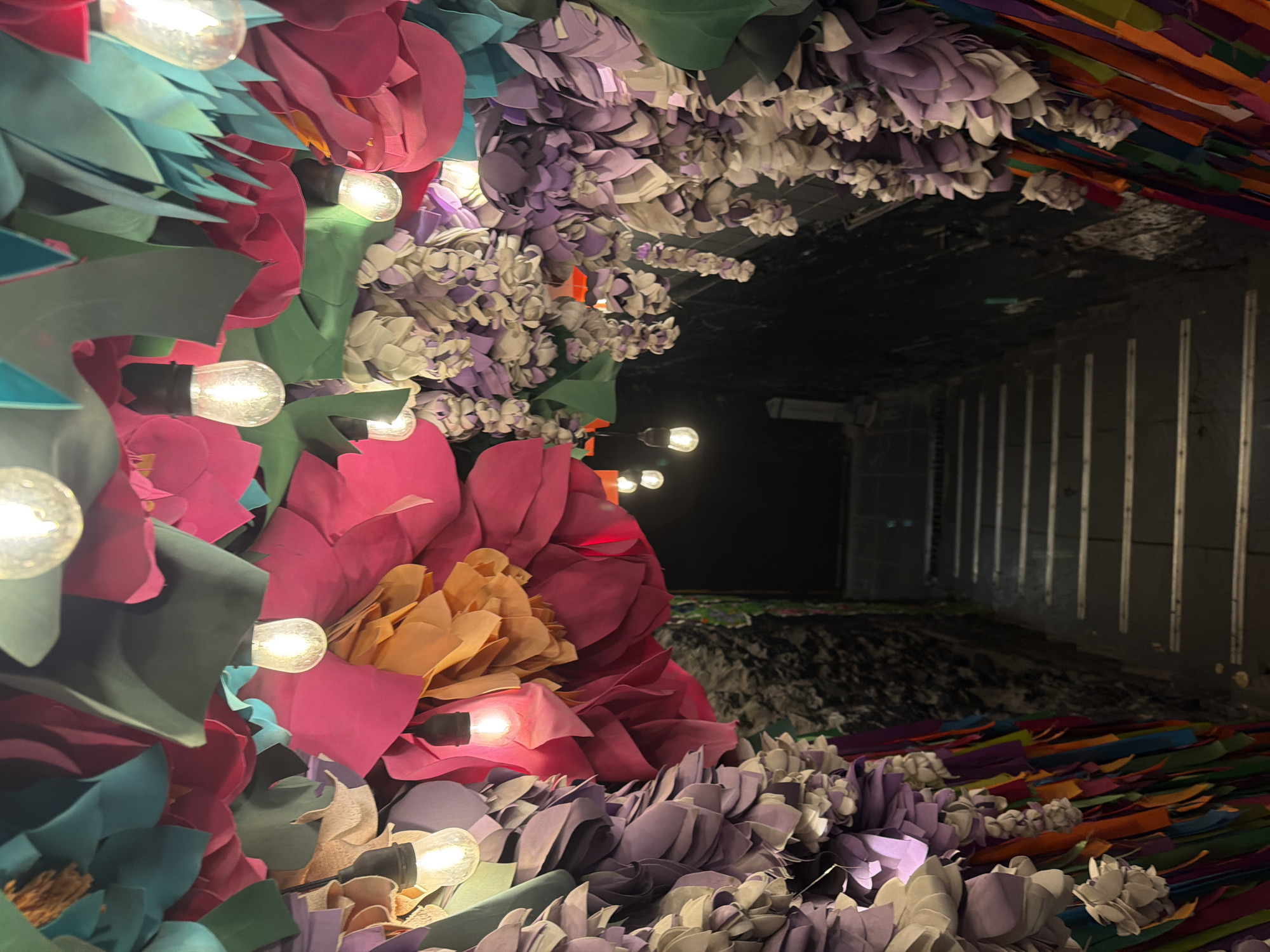 The image depicts a decorated tunnel in Rostov-on-Don, Russia. The tunnel entrance is adorned with oversized artificial flowers in shades of pink, purple, and turquoise. Strings of incandescent bulbs are interspersed among the flowers. The tunnel interior appears dark and somewhat industrial, with vertical stripes along the walls. A scattering of colorful paper strips is visible along the tunnel floor.
