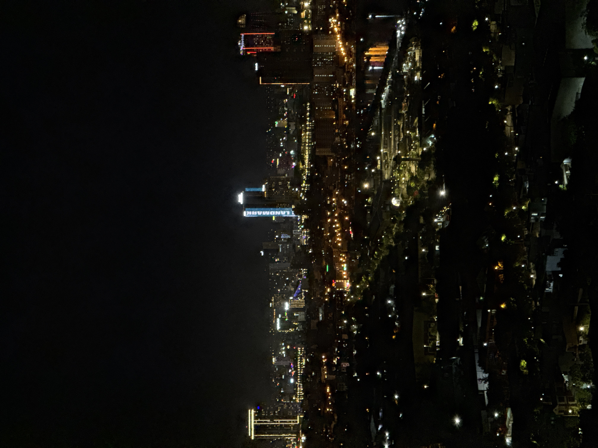The image is a night-time aerial view of the city of Đà Nẵng, Vietnam. The scene is dominated by the dark night sky and the illuminated cityscape below. Numerous buildings of varying heights are visible, many of them skyscrapers, adorned with bright lights that create a vibrant urban glow. Some buildings are lit with white, yellow, and red lights, forming distinctive patterns. The prominent "LANDMARK" sign is visible on a tall building, suggesting it is part of a business or residential complex. There is a body of water, likely a river or bay, reflecting the city lights in the middle ground. The foreground consists of darker, less illuminated areas, possibly residential neighborhoods or undeveloped land, with scattered streetlights and building lights. The overall impression is of a bustling, modern city at night.