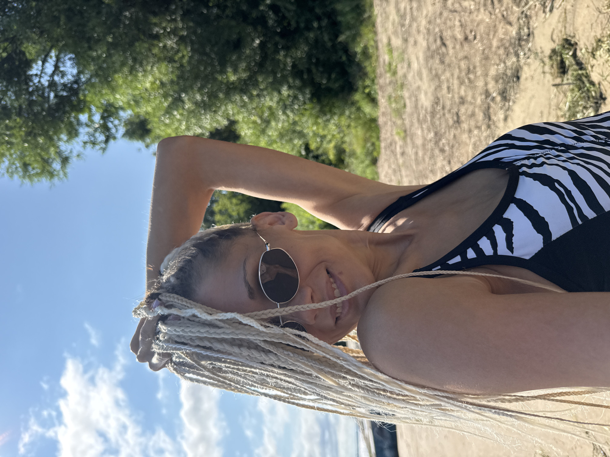 A woman with blonde braided hair and sunglasses is standing on a sandy beach, with trees and a blue sky with clouds in the background. She is wearing a black and white zebra-print swimsuit. Her left arm is raised, and she is holding her hair with her left hand. She is smiling at the camera. The location appears to be a natural outdoor setting, consistent with a beach area in or near the city of Dumino, Russia.