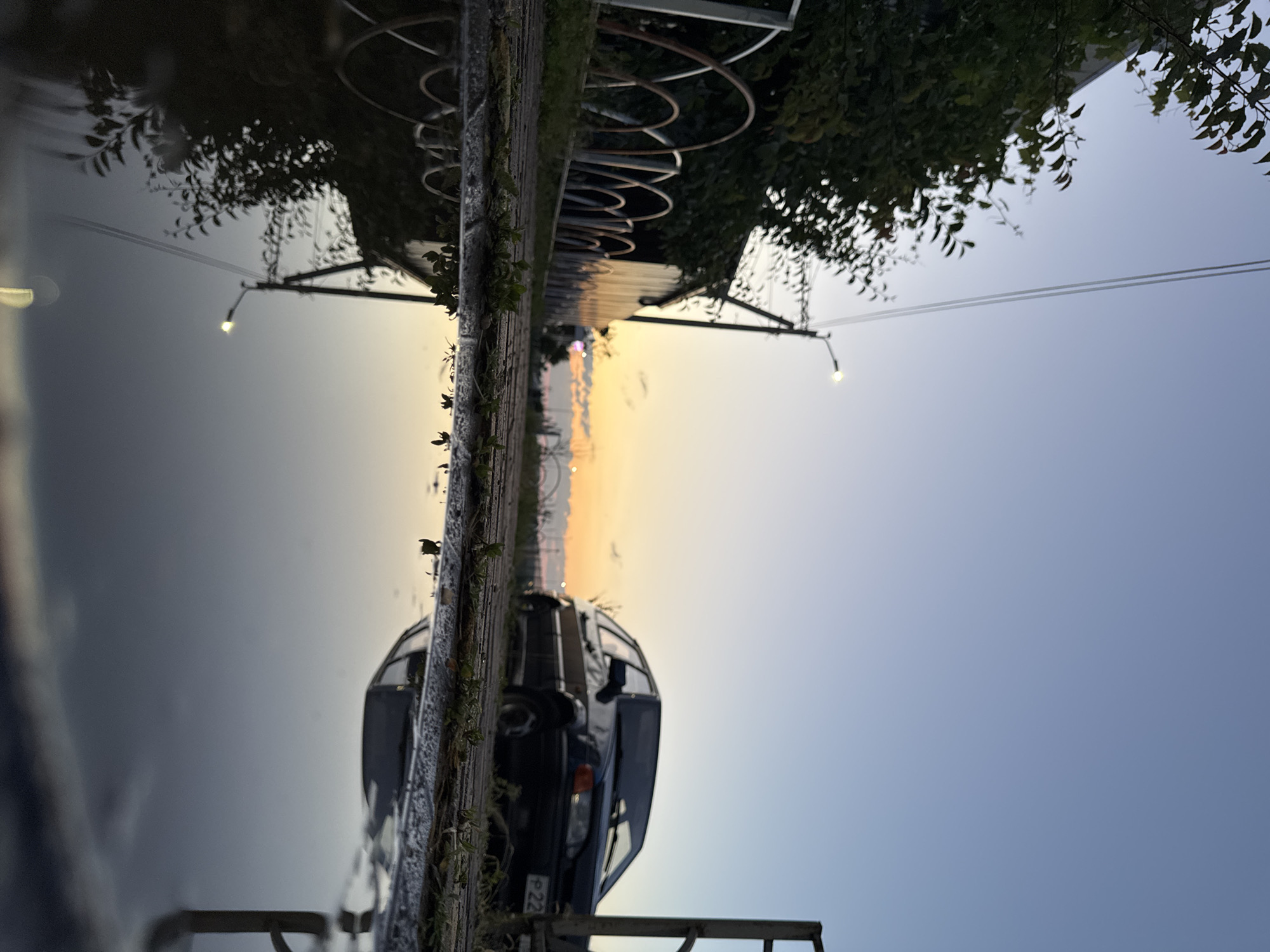 The image depicts an outdoor scene at dusk, possibly in Mikhaylovsk, Russia. The foreground is dominated by a puddle reflecting the surroundings, including a streetlamp and foliage. A black car is parked on a paved surface in the middle ground. Behind the car, a building with a corrugated metal facade is visible, partially obscured by trees and a decorative fence. The sky transitions from a pale blue to shades of orange and yellow near the horizon, indicating the sunset. The overall atmosphere is tranquil and somewhat desolate.