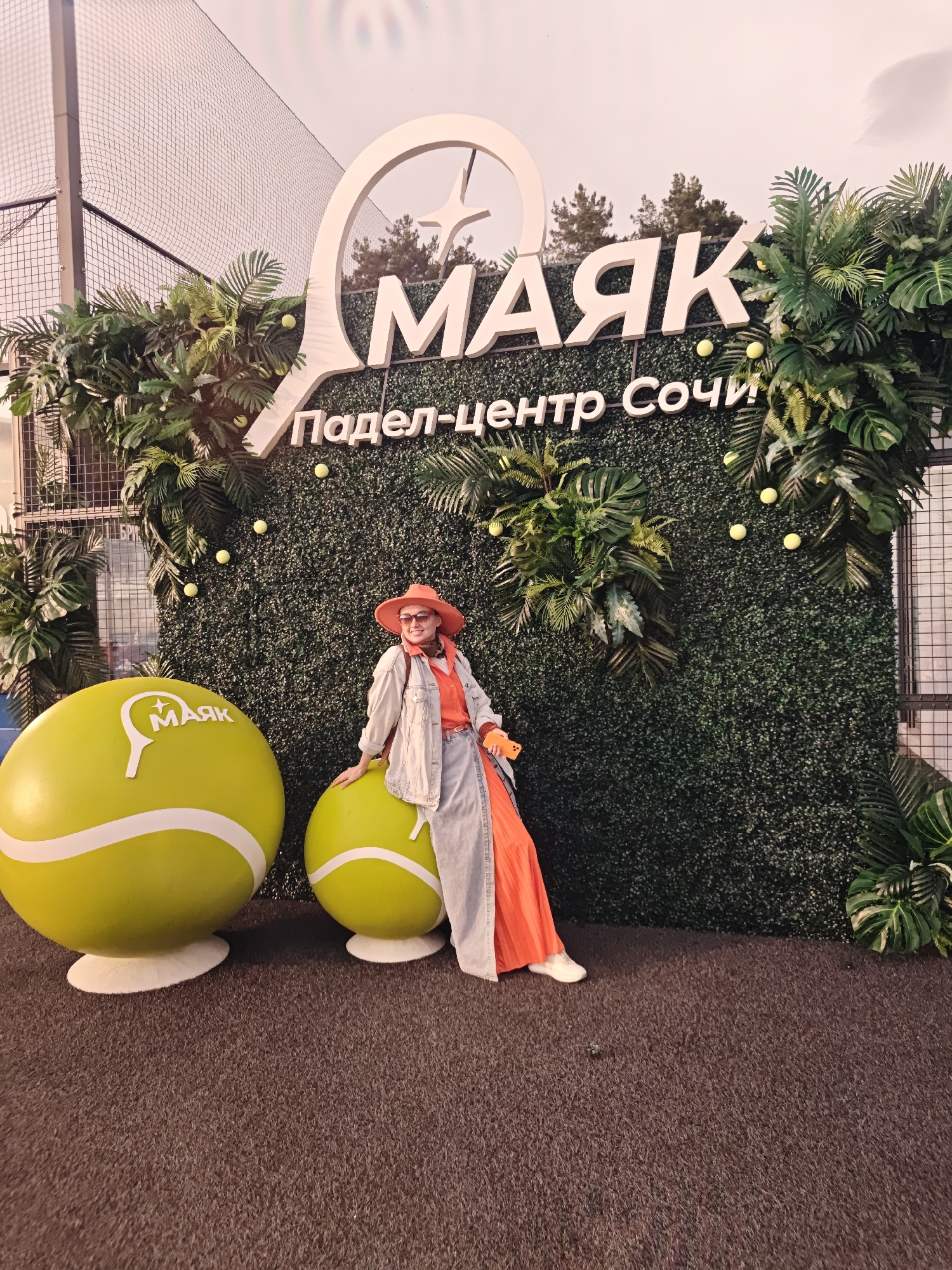 A full-shot photograph captures a woman posing in front of an advertisement for a Padel Center in Sochi, Russia.
The woman stands to the right, leaning against a large, lime-green tennis ball. She is wearing an orange hat, a matching orange top and flowing pants, a denim jacket, and white sneakers.
Behind her is a green hedge wall with tropical plants and tennis balls attached. On top of the wall is the logo of the Padel Center, featuring the word "МАЯК" (MAYAK) and "Падел-Центр Сочи" in Cyrillic lettering. Another large, lime-green tennis ball is visible to the left. The ground is covered in dark gray or brown carpeting. A partial view of a wire fence and trees are visible in the background.