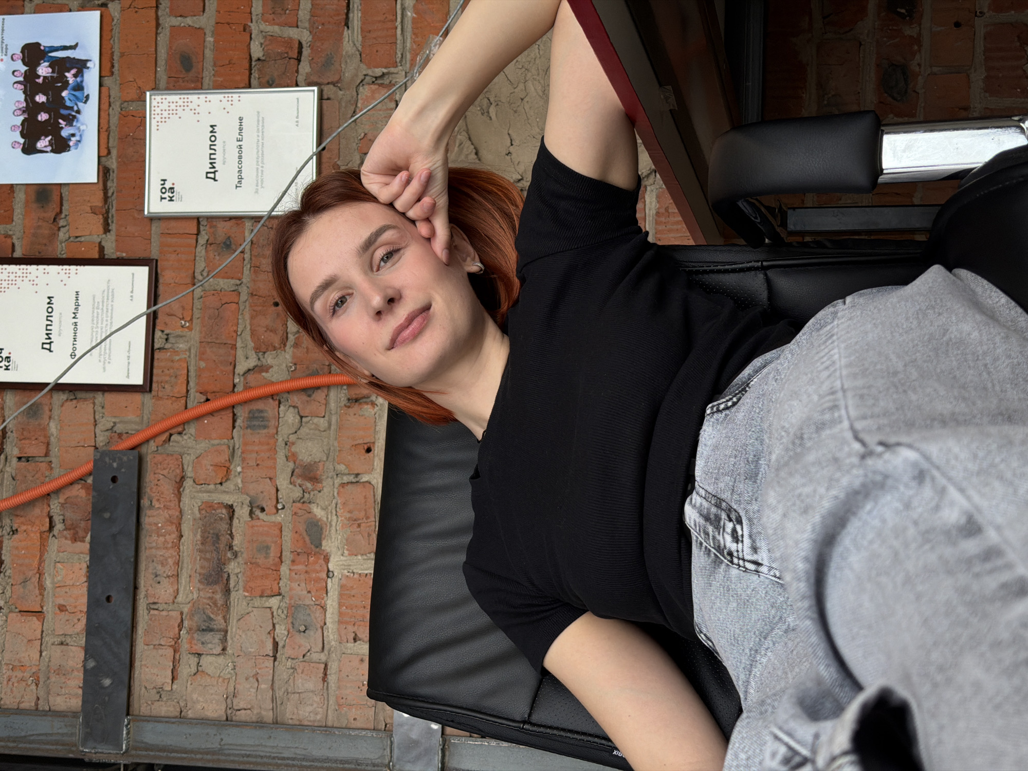 A young adult female with reddish-brown hair and light complexion is seated in a black office chair, leaning back casually. She wears a black short-sleeved ribbed top and light grey jeans. Her head is tilted slightly to the left, resting on her right hand, with a subtle smile directed towards the camera. Her left arm is down by her side, partially obscured. The chair features a textured backrest and armrests. The background consists of an exposed red brick wall, characteristic of an industrial or loft-style interior.
Affixed to the brick wall are two framed certificates and one unframed group photograph. Both certificates display the Cyrillic logo "то чка." (Tochka.) and are awards presented to "Фотиной Марии" and "Тарасовой Елене" respectively. The group photograph, positioned above the lower certificate, depicts approximately twelve individuals and is labeled as "-инструкторская группа" (instructor group). A grey metal structural beam is visible on the far left, with a black rectangular metal plate attached to it and the brick wall. An orange corrugated electrical conduit runs diagonally across the brick wall, starting from the upper left and moving towards the lower right. The upper edge of a dark red surface, likely a desk or table, is visible on the right side of the image, beneath the woman's right arm.
Based on the visible "то чка." (Tochka.) branding and the Russian language text, this scene is located within "Точка Кипения Чайковский" (Boiling Point Chaikovsky), an innovation space in the city of Chaikovsky, Russia.