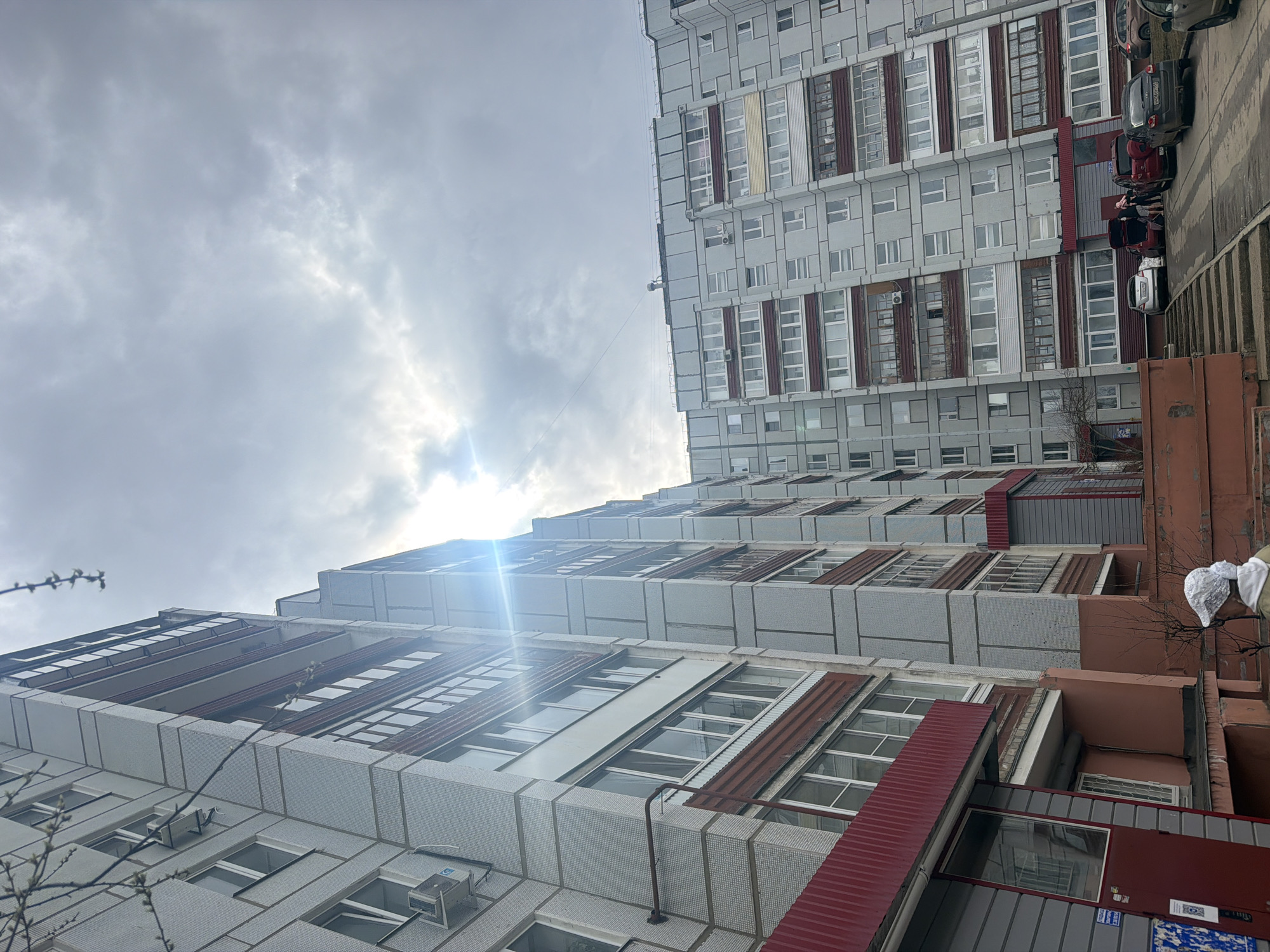 The image shows the exterior of a high-rise building complex in Chaikovsky, Russia. The building's facade is predominantly grey and features a modular design with rows of windows. Some windows have visible damage. Maroon colored accents are integrated into the building's exterior design. The sky is overcast. Several cars are visible to the right of the structure. The photograph is taken from a low-angle perspective, looking upwards.