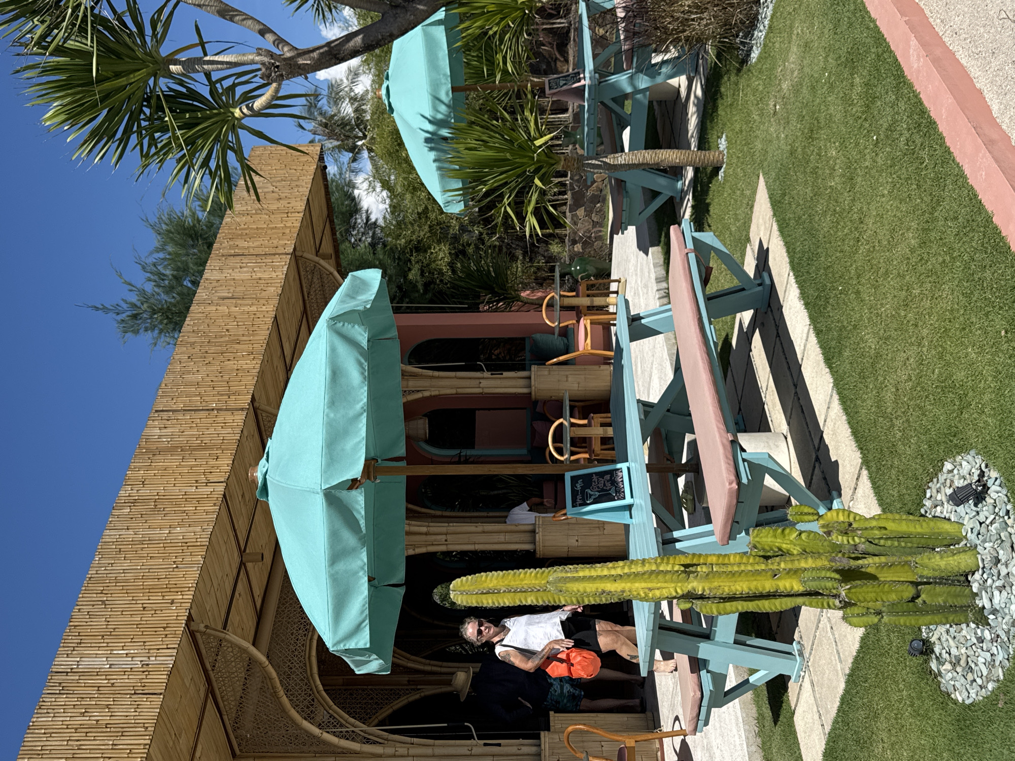 A man wearing a white shirt and shorts is standing near a cactus and a blue picnic table with an umbrella in what appears to be a restaurant or outdoor area in the city of Unknown, Indonesia. The area is decorated with bamboo structures and turquoise umbrellas, and there are additional tables and chairs in the background. The surrounding environment consists of green grass and trees.