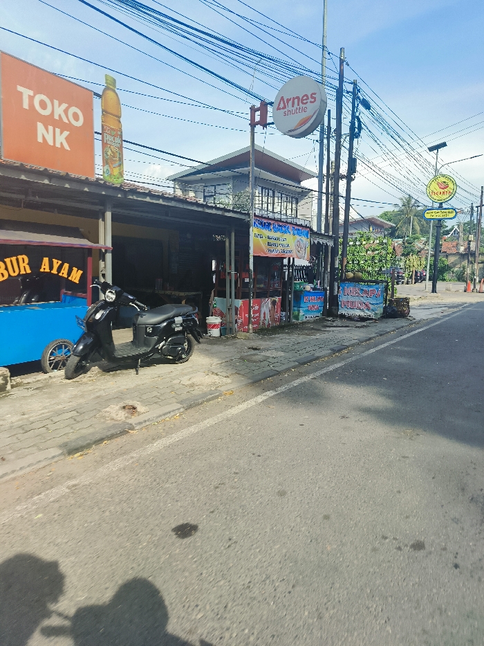 The image shows a street scene in Jatiluhur, Indonesia, at the coordinates -6.565 latitude and 107.434 longitude. The scene is captured from a street-level perspective, looking towards the end of a paved road. On the left side of the image, there is a row of storefronts. The closest storefront is marked with a sign that says "TOKO NK" in large, bold letters, while the next storefront displays a sign that reads "BUR AYAM." There are numerous electrical wires running overhead. A black scooter is parked on the sidewalk in front of these storefronts, with its front wheel facing the street. Several other objects can be seen on the sidewalk, including a blue cart with a black wheel and a metal bucket. The street itself is paved and appears to be relatively empty, with only a few shadows cast by buildings and trees. There are trees and shrubs lining the side of the road, and a few signs, including one that reads "Arnes shuttle." The image suggests a casual, everyday scene in a bustling city, with a mix of commercial activity and residential elements.