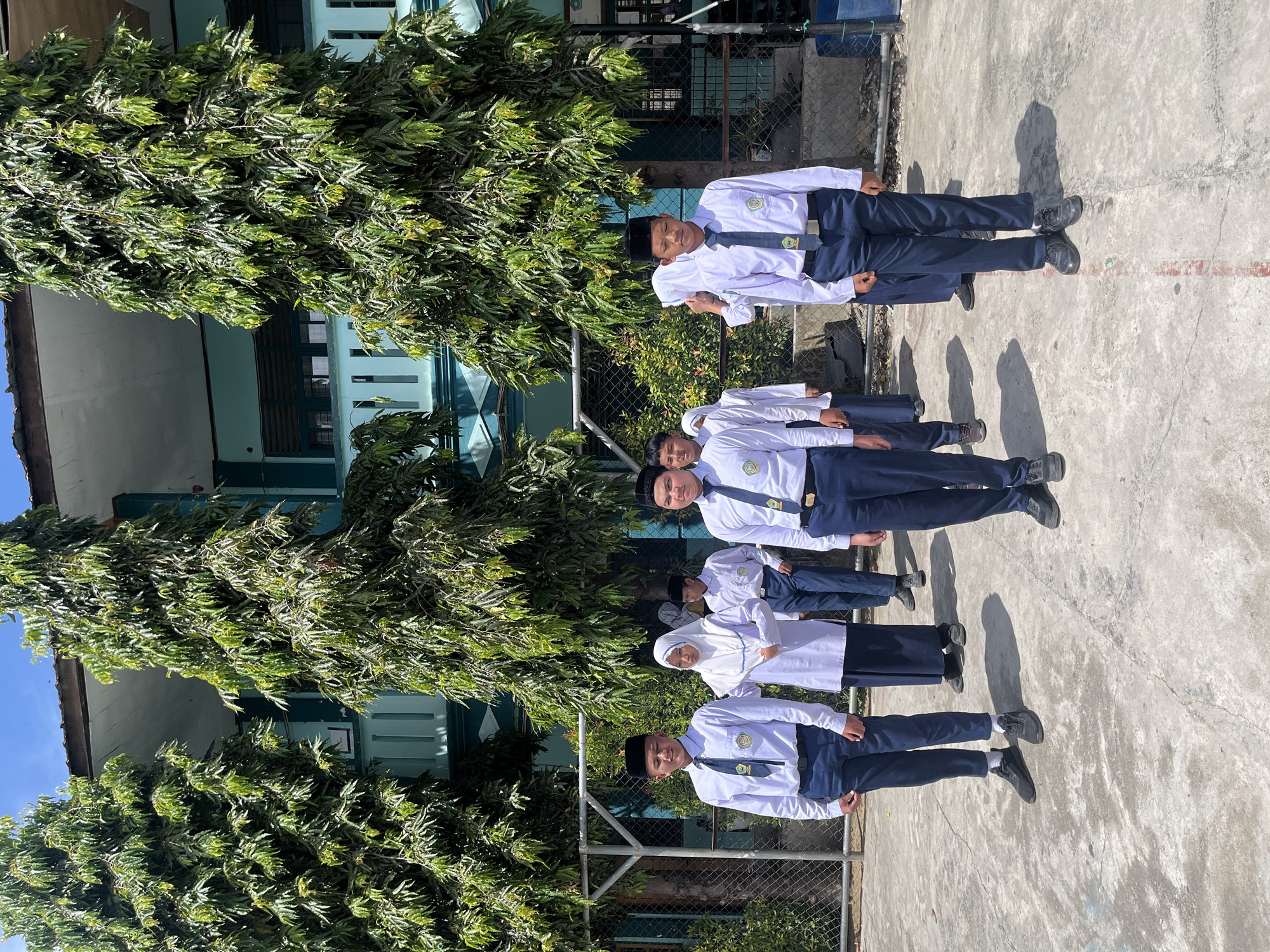 A group of students, a mix of boys and girls, stand in formation outdoors. The boys are wearing white long-sleeved shirts, dark blue pants, black peci hats, and dark ties. The girls are wearing white headscarves and white long-sleeved tops with dark blue skirts. They are standing on a paved area in front of a two-story building with a green facade and large trees with green foliage. The building appears to be a school. The scene is well-lit by sunlight, casting shadows on the ground. This image was taken at a school in Lhokseumawe, Indonesia.