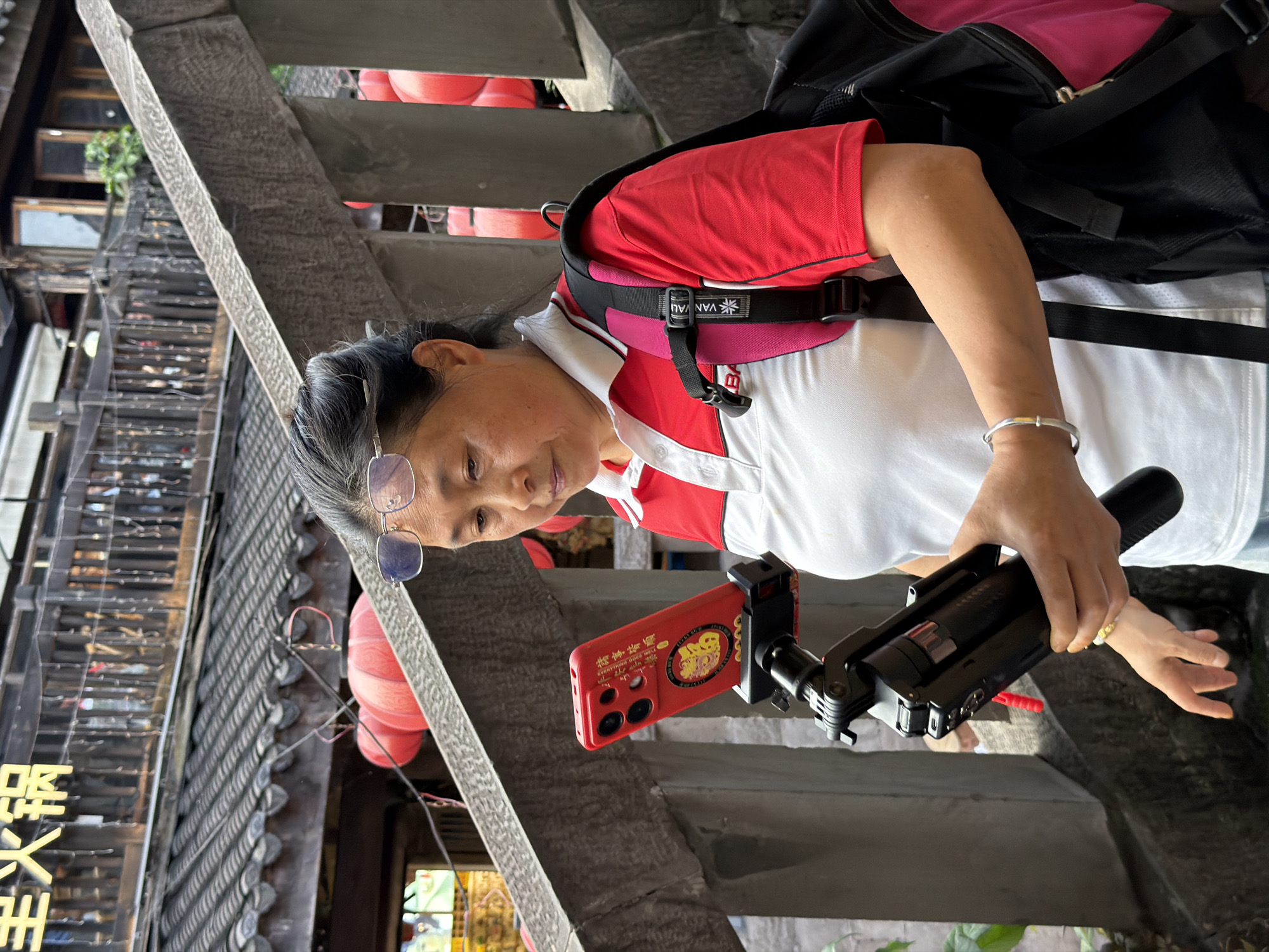 A medium shot captures an East Asian woman in Yuzhong District, China, standing on a stone pathway with a backpack. She wears glasses, a red and white polo shirt, and holds a smartphone mounted on a black tripod in her hands. The phone is red and has a decorative case. Behind her, traditional Chinese architecture with tiled roofs and red lanterns are visible. The woman appears to be looking at her phone. The scene suggests an outdoor exploration or recording activity in a culturally rich environment.
