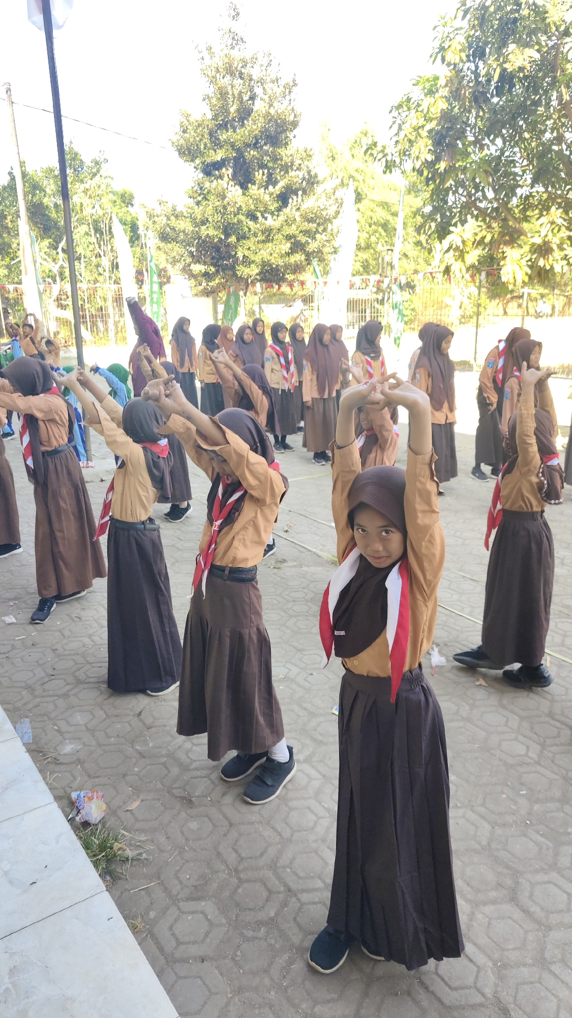 The image shows a group of young girls, likely students, performing an outdoor physical activity. They are dressed in uniforms consisting of light brown shirts, dark brown skirts, and hijabs, with some wearing red and white scarves around their necks, indicating a scout or similar organizational affiliation. The girls are arranged in rows and appear to be stretching or doing calisthenics, with many of them raising their arms. The setting is an outdoor paved area with a textured surface, possibly a schoolyard or public space. In the background, there are trees with lush green foliage, flags, and what appears to be a fence. The overall scene suggests a structured group activity taking place in a warm, possibly sunny, environment. The image quality is somewhat grainy, and the lighting is bright, suggesting it was taken during the daytime.