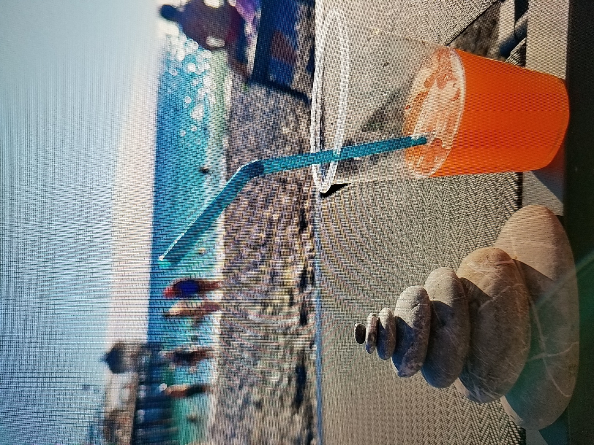 The image depicts a beach scene with a stacked stone cairn and a plastic cup of orange liquid with a blue straw in the foreground. In the background, the ocean and a pier can be seen. Several people are visible near the water's edge. The setting appears to be a sunny day. The location is not Tiraspol, Moldova; it is a beach.