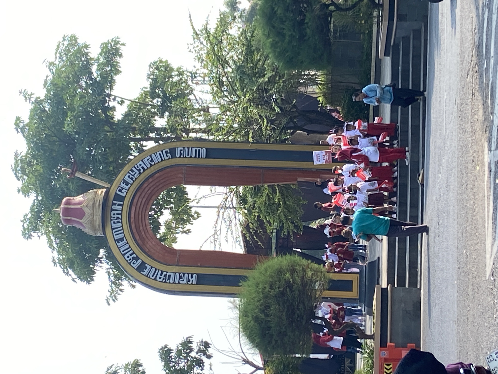 In the city of Kecamatan Pati, Indonesia, a group of children in red and white uniforms are gathered near a large archway. The archway features text in an Indonesian language and is topped with a crown-like structure. The children are holding small Indonesian flags and appear to be participating in a parade or event. A man in a teal shirt is walking away from the group, carrying a child. Another woman in a blue jacket is walking down a set of stairs. The background consists of trees and a bright, overcast sky. The scene is set on a paved area with a road in the foreground.