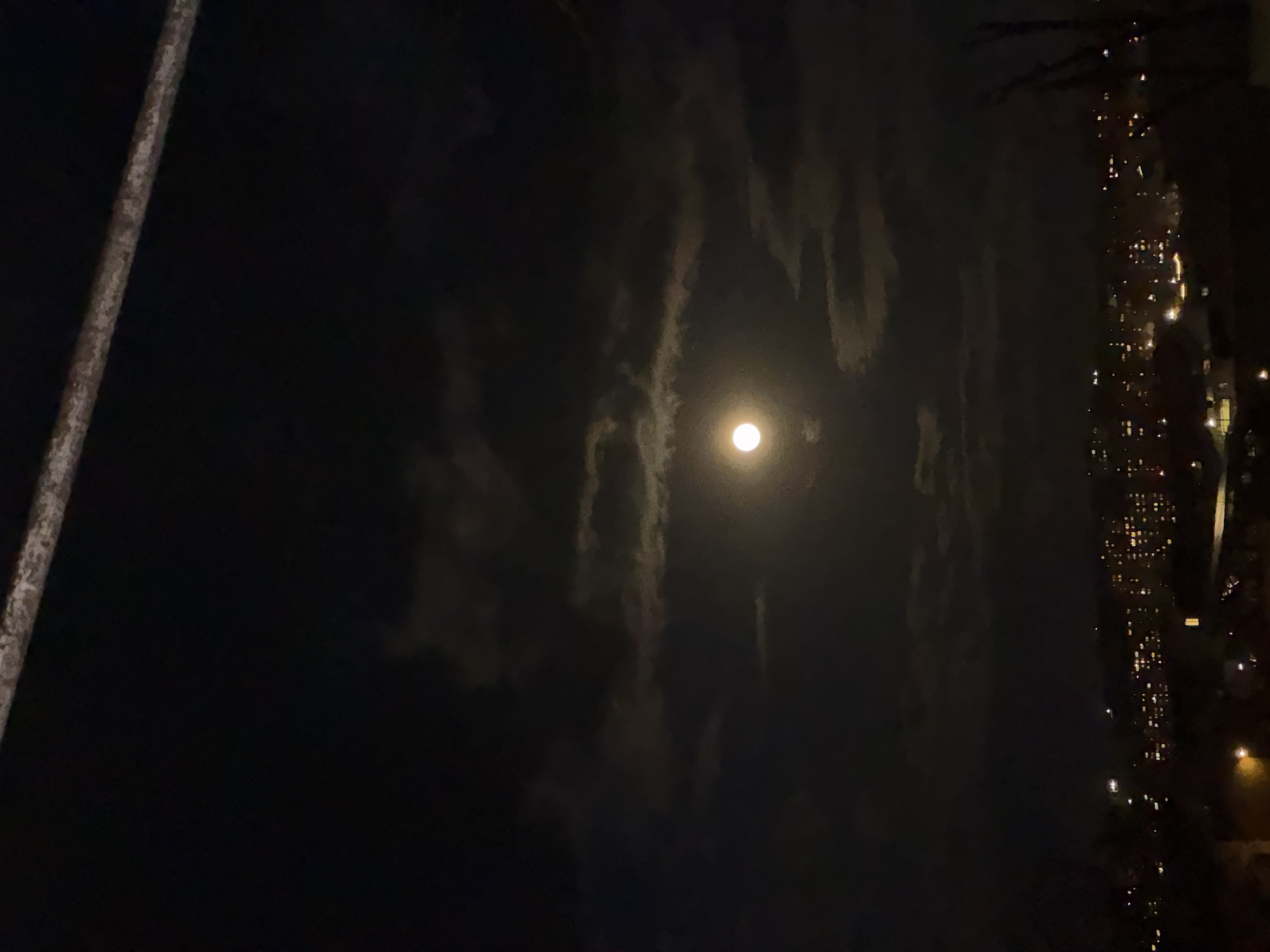 The image shows a night scene in Yerevan, Armenia. A full moon is visible in the center of the frame, partially obscured by wisps of clouds. To the left, a pole-like structure is present. On the right, illuminated windows suggest a dense urban environment. The rest of the picture is a dark scene. There is no evidence in the frame that reveals the presence of any people or their actions.