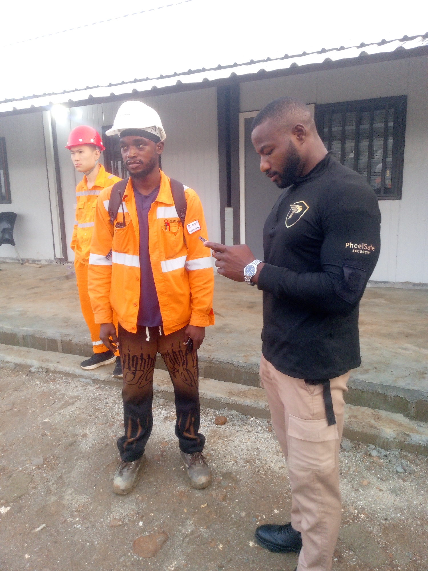 In Indo, Nigeria, three men are standing in front of a white building with a corrugated roof. The man on the left wears a red hard hat and an orange jumpsuit with reflective stripes. The middle man wears a white hard hat and an orange jacket with reflective stripes over a purple shirt, with dark pants featuring a decorative pattern. He is looking towards the right. The man on the right wears a black long-sleeved shirt with a gold emblem on the chest and "PheelSafe SECURITY" printed on the sleeve, and light brown cargo pants. He is looking down at his phone, which he is holding in his right hand, and he is wearing a wristwatch on his left wrist. The ground is covered in dirt and gravel.