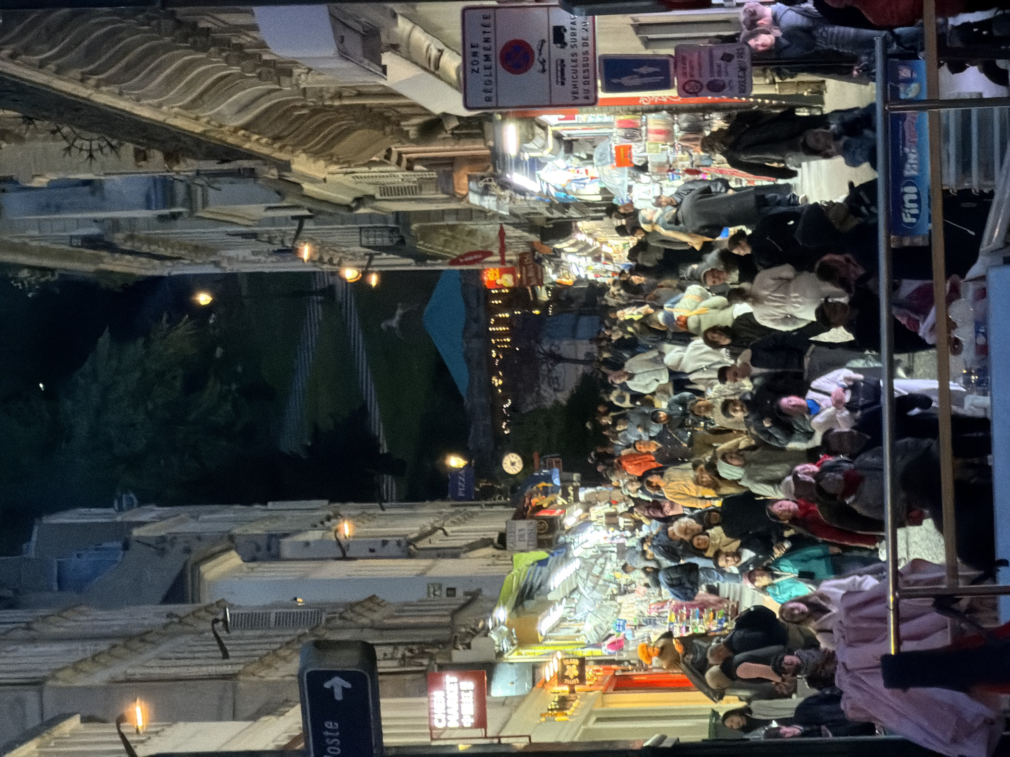 Here's a description of the image:
The image captures a bustling scene on a Parisian street at night. A crowd of people, densely packed, walks through a market area. Stalls are lit up, displaying various wares, including food items and other goods. Above the scene, street signs are visible, one indicating parking regulations and another pointing to a post office. Architectural details of Parisian buildings line the street, with ornate facades and lit windows. The image suggests a vibrant, crowded urban environment with a mix of residents and tourists.