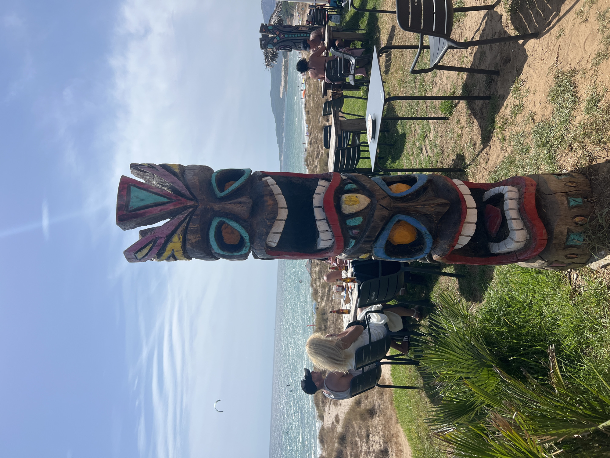 A colorful, carved tiki statue stands prominently in the foreground, its wooden form adorned with vibrant paints and featuring a wide, open mouth with visible teeth and tongue. Behind the statue, a beachside restaurant is in view. Several wooden tables and black chairs are set up on a grassy area, with some occupied by patrons. In the mid-ground, people are seated at tables, some appearing to be eating or drinking, while others are conversing. Bottles and plates are visible on the tables. Beyond the restaurant seating, a sandy beach stretches out, meeting the sparkling blue ocean. Gentle waves roll onto the shore, and a few people are visible on the beach, some possibly swimming or sunbathing. In the distance, a kiteboarder is visible on the water, and hills form the backdrop along the coastline. The sky is a clear blue with scattered, wispy clouds. The overall scene depicts a relaxed, outdoor dining experience by the sea in Tarifa, Spain.