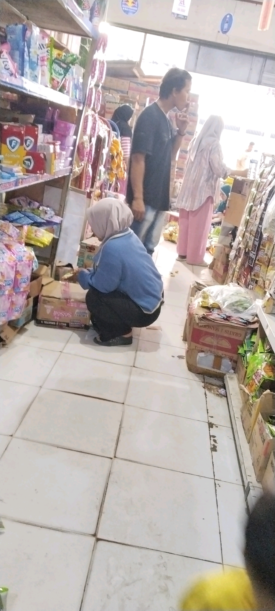 The image depicts the interior of a convenience store, possibly in Bandar Jaya, Indonesia, with several individuals browsing or working. In the foreground, a person wearing a blue sweatshirt and a light-colored hijab is squatting down, examining items in a cardboard box on the tiled floor. To the left, shelves are stocked with various packaged goods, including brightly colored bags and boxes with prominent branding. In the mid-ground, a man in a dark t-shirt stands with his head slightly bowed, and further back, another person in a light-colored striped top and pink pants is visible. The store appears to be well-lit, with natural light entering from the right side of the frame, creating bright highlights and some glare on the products. Cardboard boxes are stacked on the floor and on shelves, and various items, some in plastic packaging, are displayed throughout the store. The overall impression is of a busy retail environment with diverse products for sale.