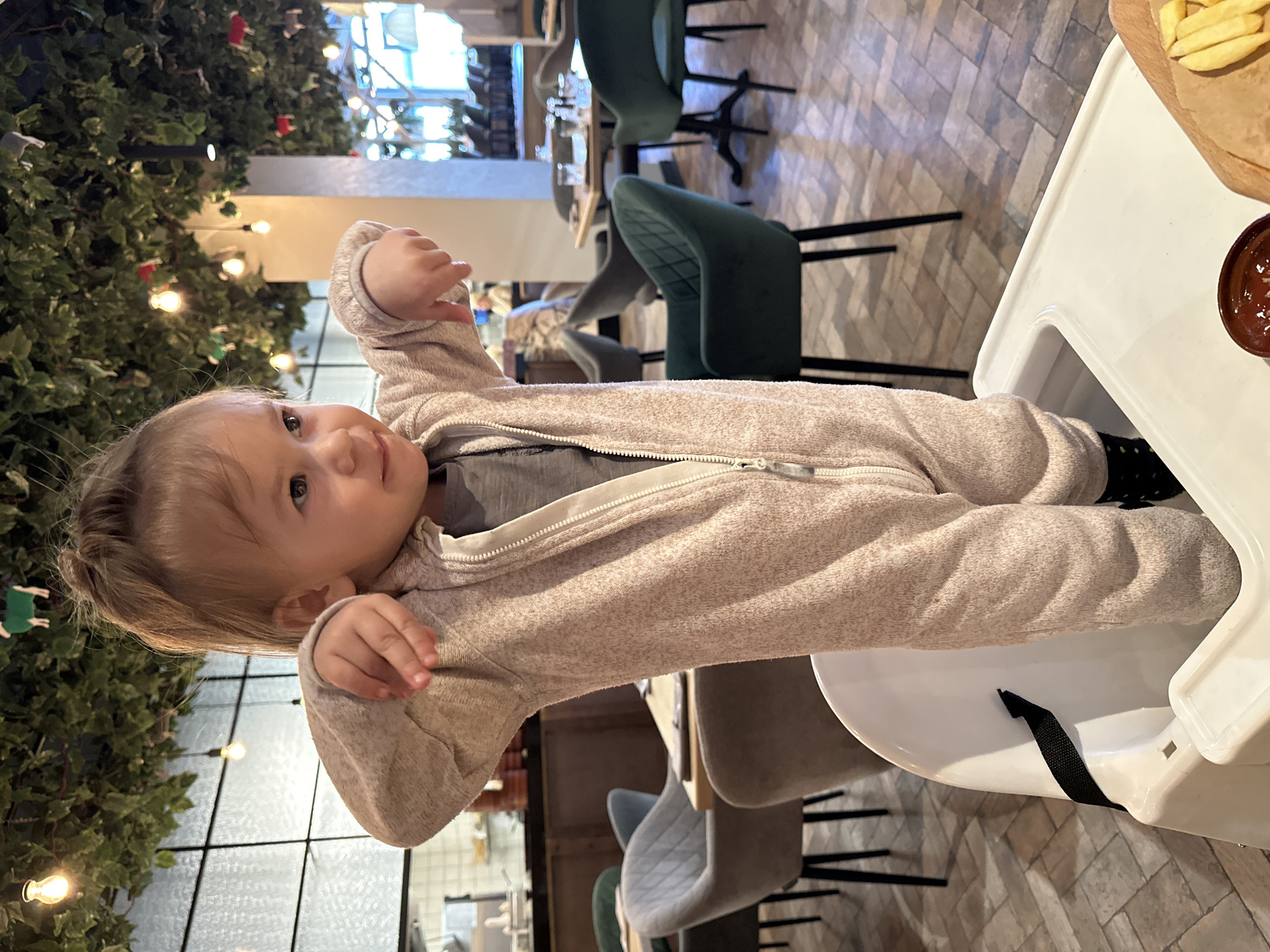 A toddler in a beige, zipped-up jumpsuit sits in a white highchair in what appears to be a restaurant. On the highchair's tray are french fries and a small bowl of sauce. The restaurant has green chairs and a decorative green wall with lights. There is no information to indicate this image was taken in the city of Izhevsk, Russia.