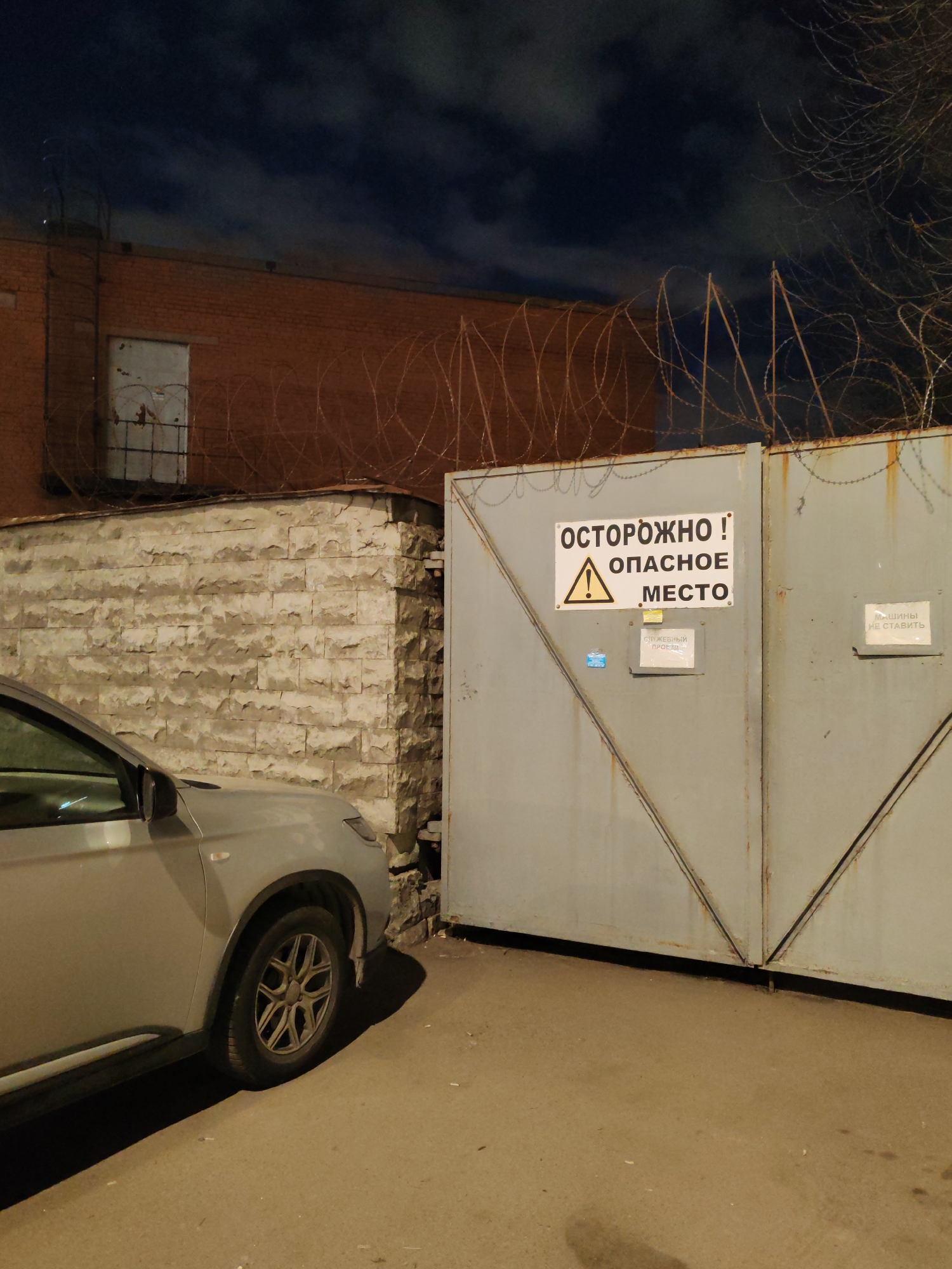 The image captures a nighttime scene in what appears to be an industrial or restricted urban area in Saint Petersburg, Russia. The primary subjects are a light-colored car, a large metal gate, a stone wall, and a brick building, all under a dark, cloudy night sky.
On the left, the front portion of a light grey car, possibly an SUV or crossover, is parked on a paved surface. Its front wheel and a significant part of its side are visible, illuminated by an unseen light source that casts long shadows to the right. To the right of the car is a textured stone wall, built from rough-hewn blocks, which forms a corner and extends towards the large gate. Behind this wall and above it, a red-brick building is visible, with a boarded-up or closed window on its side. Barbed wire is extensively strung along the top of the brick building and across the top of the metal gate, suggesting a secure or high-security area.
The prominent feature is a large, grey, double-leaf metal gate with diagonal bracing. Several signs are affixed to it. A large warning sign in the center reads "ОСТОРОЖНО! ОПАСНОЕ МЕСТО" (CAUTION! DANGEROUS PLACE) with a yellow warning triangle icon. Below it, a smaller sign indicates "СЛУЖЕБНЫЙ ПРОЕЗД" (SERVICE DRIVEWAY or STAFF ACCESS). On the right panel of the gate, another sign states "МАШИНЫ НЕ СТАВИТЬ" (DO NOT PARK CARS or NO PARKING). The presence of these signs, coupled with the barbed wire, strongly indicates a restricted access zone. The sky is dark with visible clouds, and the scene is lit by artificial lights, creating strong contrasts and shadows. No human activity is depicted.
