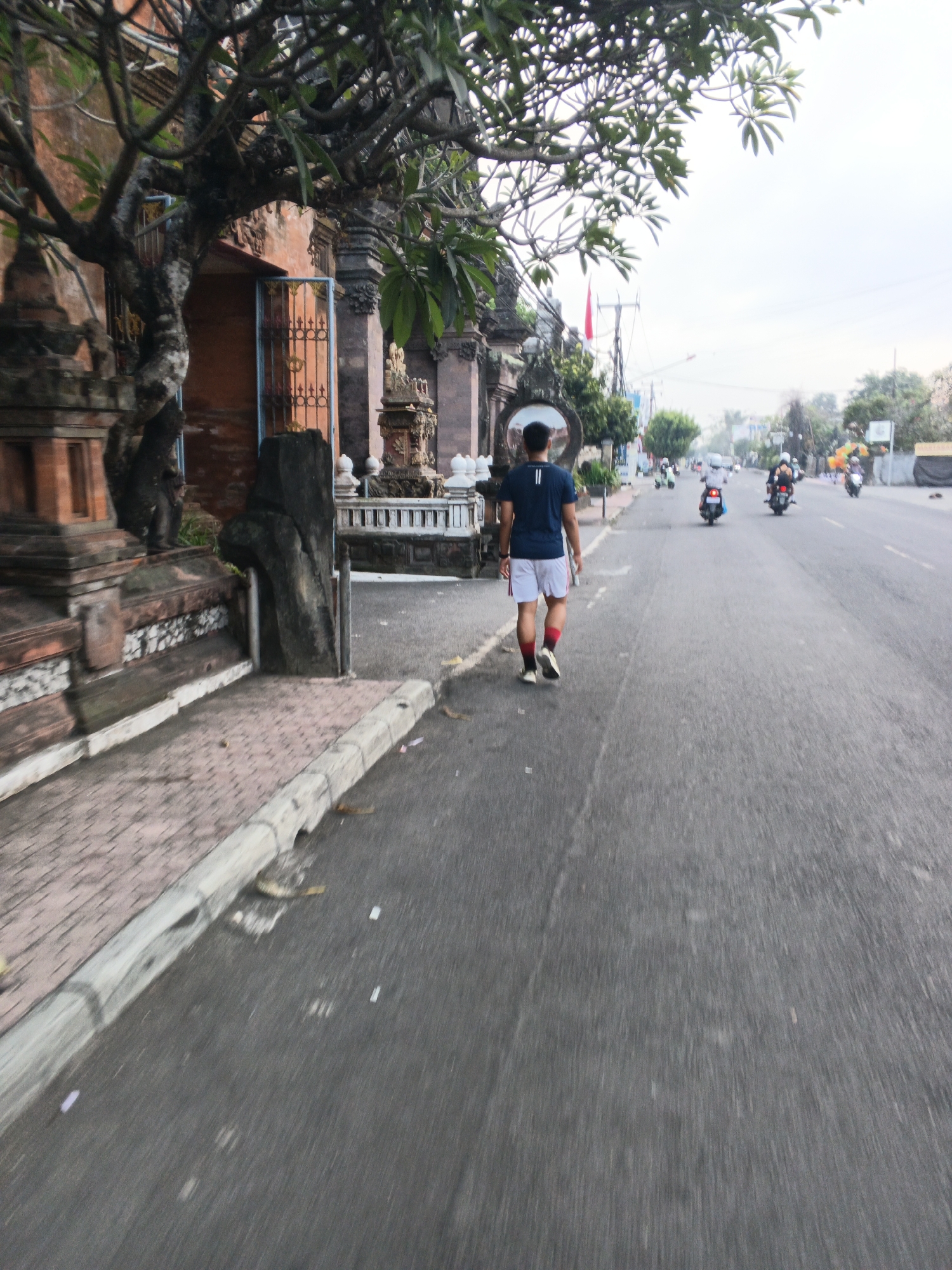 A person walks away from the camera on a paved road in Sukawati, Indonesia, on a cloudy day. The person is wearing a dark blue t-shirt, white shorts with red accents, and red and white socks with black shoes. To the left of the road is a sidewalk with a red-tiled pattern and a low stone wall. Adjacent to the sidewalk is a building with traditional Balinese architecture, featuring ornate stonework, a terracotta-colored facade, and intricate metal gates. A large tree with dark branches and green leaves overhangs the building and sidewalk. Beyond the building, there is a street with several motorcycles and scooters in motion. The road appears to be a main thoroughfare in the town, with shops and other buildings visible in the distance. The overall impression is of a typical street scene in a Southeast Asian town.