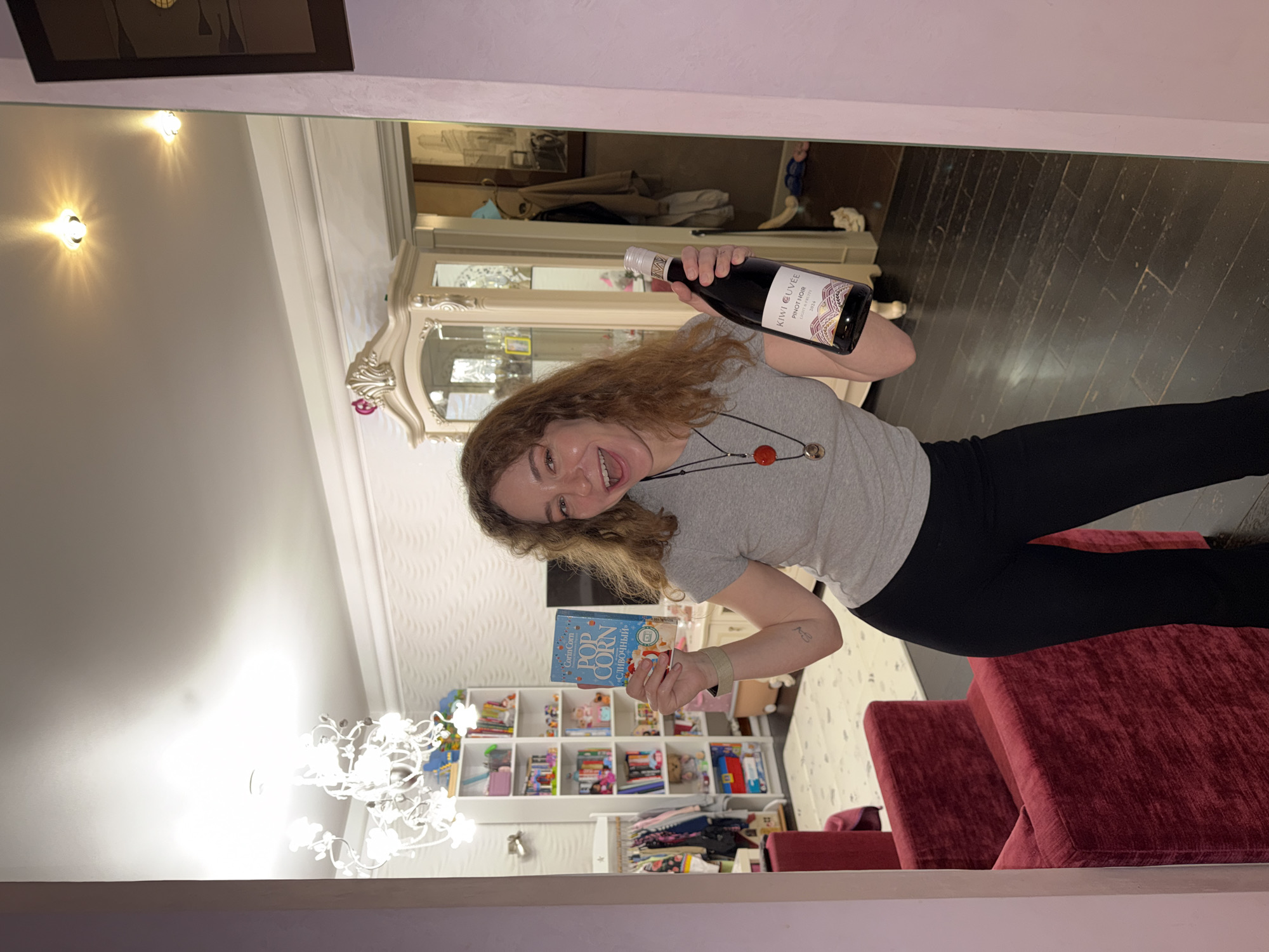 A woman stands in front of a mirror, holding a bottle of Kiwi Cuvée Pinot Noir and a box of Corin Corn Popcorn "СЛИВОЧНЫЙ" (creamy) in what appears to be an apartment. She is wearing a gray t-shirt, black leggings, and a necklace with red accents. The apartment features a white cabinet, a bookshelf filled with colorful items, a chandelier, and burgundy upholstered furniture. The city mentioned, Moscow, Russia, cannot be verified from this image alone.