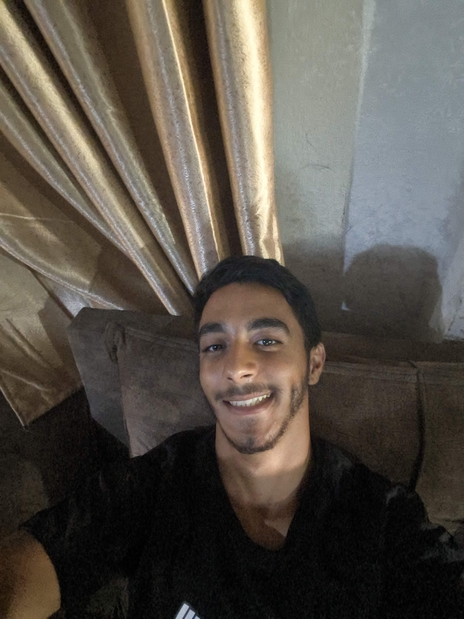 A young man with dark hair, a short beard, and brown eyes smiles at the camera. He is wearing a black t-shirt. He is sitting on a brown couch in front of a gold curtain and a textured wall. The lighting in the room is dim, casting shadows on the wall behind him. The image appears to be a selfie.