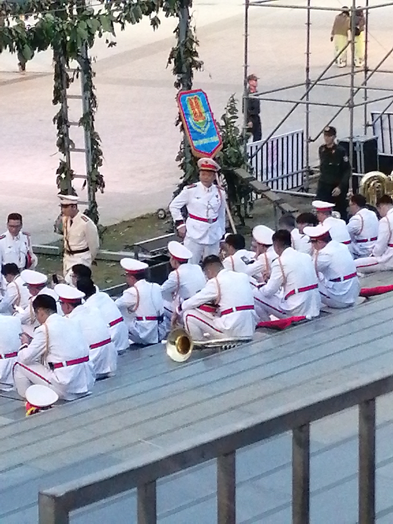 The image shows a large group of people, most of whom are dressed in white military-style uniforms with red trim. Many are seated on bleachers, and some are holding musical instruments, including a tuba. A few individuals in darker uniforms are standing, and one is holding a banner. The scene appears to be an outdoor event, possibly a parade or performance, with scaffolding and greenery in the background. The lighting suggests it is daytime. Based on the presence of similar events and architectural styles, this scene is consistent with the Da Lat Flower Festival in Da Lat, Vietnam, which often features parades and marching bands in ceremonial attire.