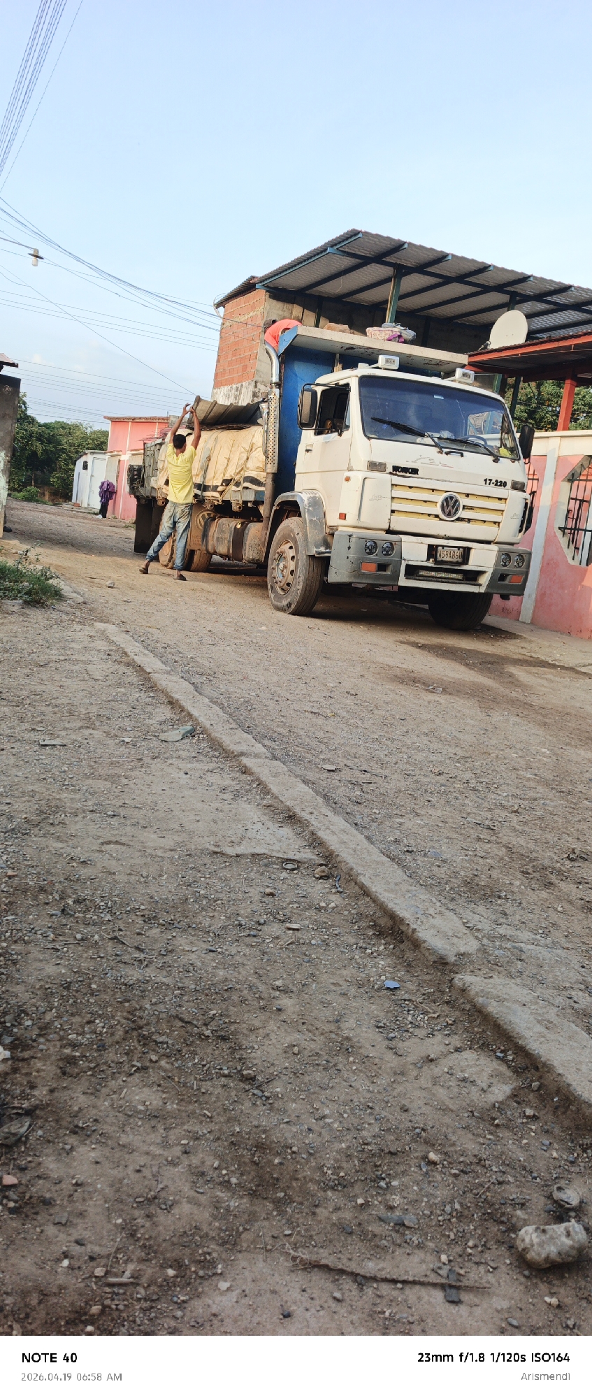 A man is standing on the back of a parked dump truck on a dirt road in the city of Arismendi, Venezuela. The truck is white with a blue dump bed covered by a tan tarp, and it is parked on the right side of the dirt road. The man is wearing a yellow shirt and blue jeans and appears to be adjusting the tarp. There is a building next to the truck on the right side of the image, with a red and white fence. In the background, there are other buildings and a person walking away from the camera in a purple dress. The foreground is composed of a dirt road with a concrete curb. The time and date stamp is visible at the bottom left corner of the image, reading "2026.04.19 06:58 AM", and the camera settings are visible at the bottom right corner of the image, reading "23mm f/1.8 1/120s ISO164 Arismendi".