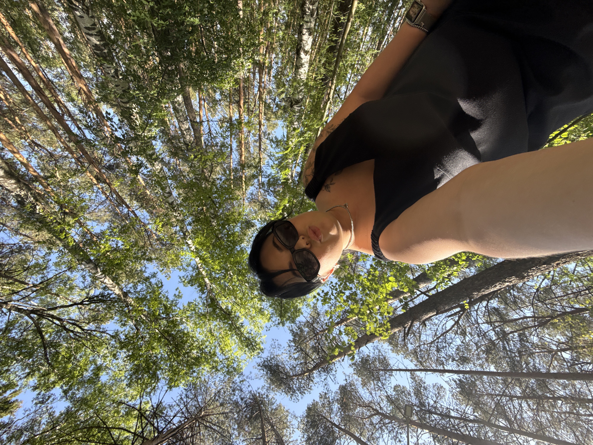 The photograph appears to have been taken in a forest in Chaikovsky, Russia. The perspective is from below, looking upwards toward the sky through the dense canopy of trees. A woman is prominently positioned in the foreground, seemingly holding the camera or phone to capture the shot. She is wearing sunglasses and a dark outfit. The trees appear to be a mix of birch and pine species, consistent with the typical boreal forests of the region. Sunlight filters through the leaves, creating a dappled effect. The sky is partially visible between the branches, suggesting a clear day.