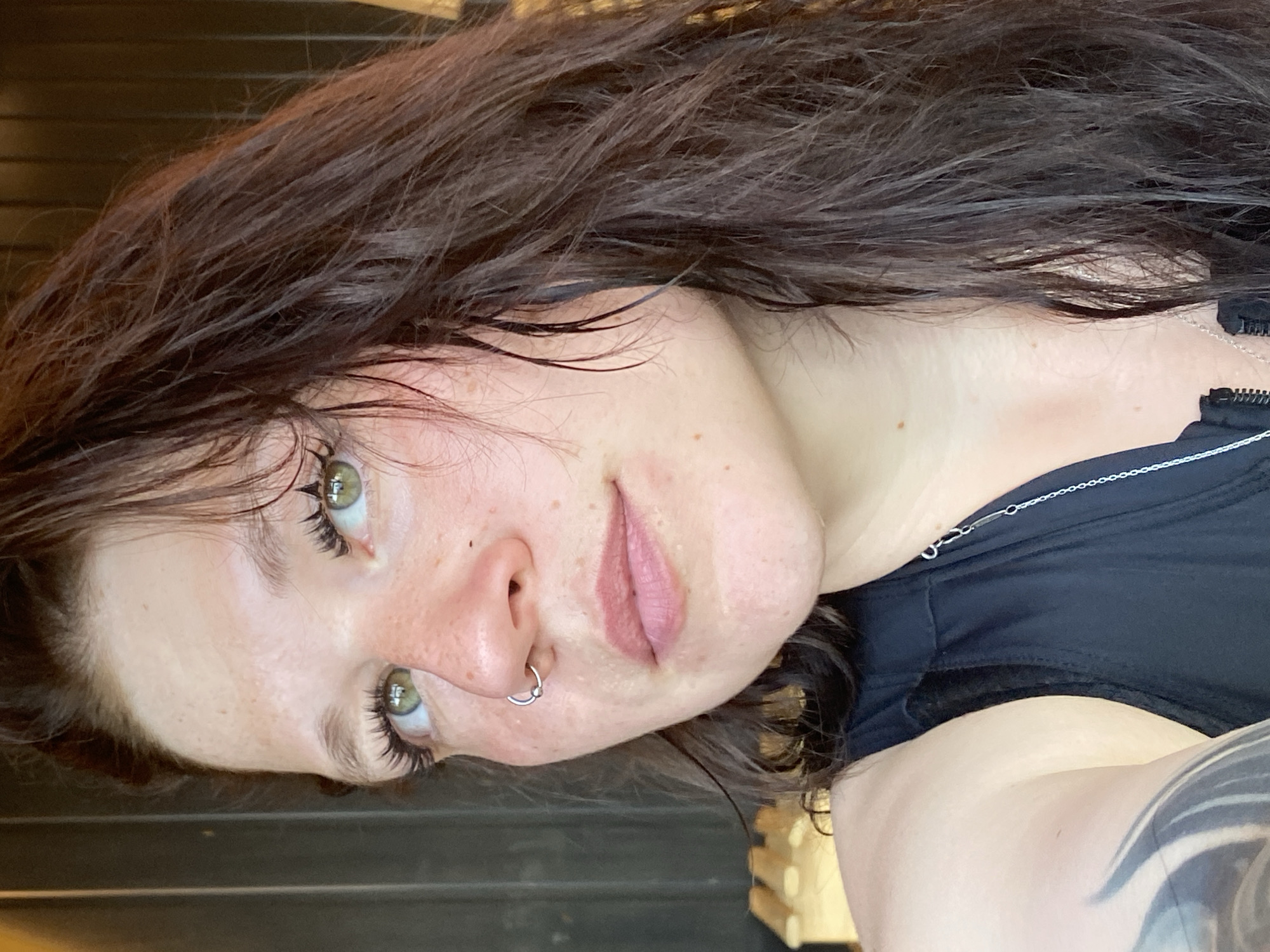 The media file is a close-up, low-angle, portrait-style photograph of a young woman. She has long, dark brown, wet hair that is styled to frame her face. Her skin is fair and appears to have some light freckles and blemishes. She has green eyes with prominent eyelashes and eyebrows. Her lips are slightly parted and appear to be a light pink color. She is wearing a silver hoop earring in her nose and a thin silver necklace. She is wearing a black garment, possibly a top or dress, with a zippered neckline. The background is dark and appears to be outdoors with some horizontal lines suggesting wooden slats or blinds. The lighting is natural and suggests it may be daytime. The woman's expression is neutral or slightly contemplative. There is no visible text in the image.