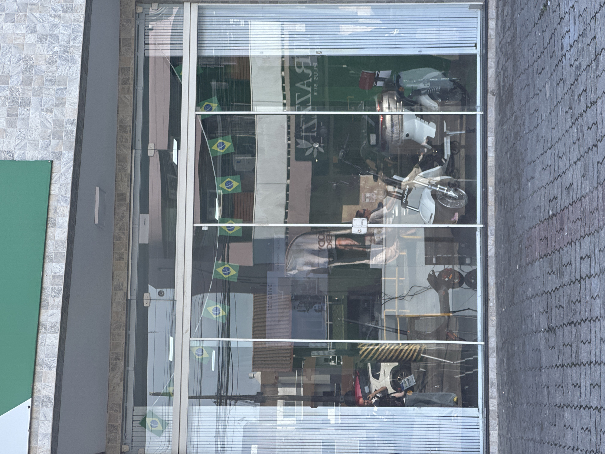 The image shows the storefront of a business in Florianópolis, Brazil, featuring a large glass facade. The facade is adorned with a string of small Brazilian flags. Inside the store, various vehicles, including a white electric scooter and parts of what appears to be a white car, are visible. A person, possibly a customer or employee, can be seen through the glass, standing near the car and holding a phone. Reflections on the glass reveal the street and surrounding buildings. The building itself has a facade clad in patterned tiles and a green awning above the storefront. The ground in front of the store is paved with dark gray interlocking stones.