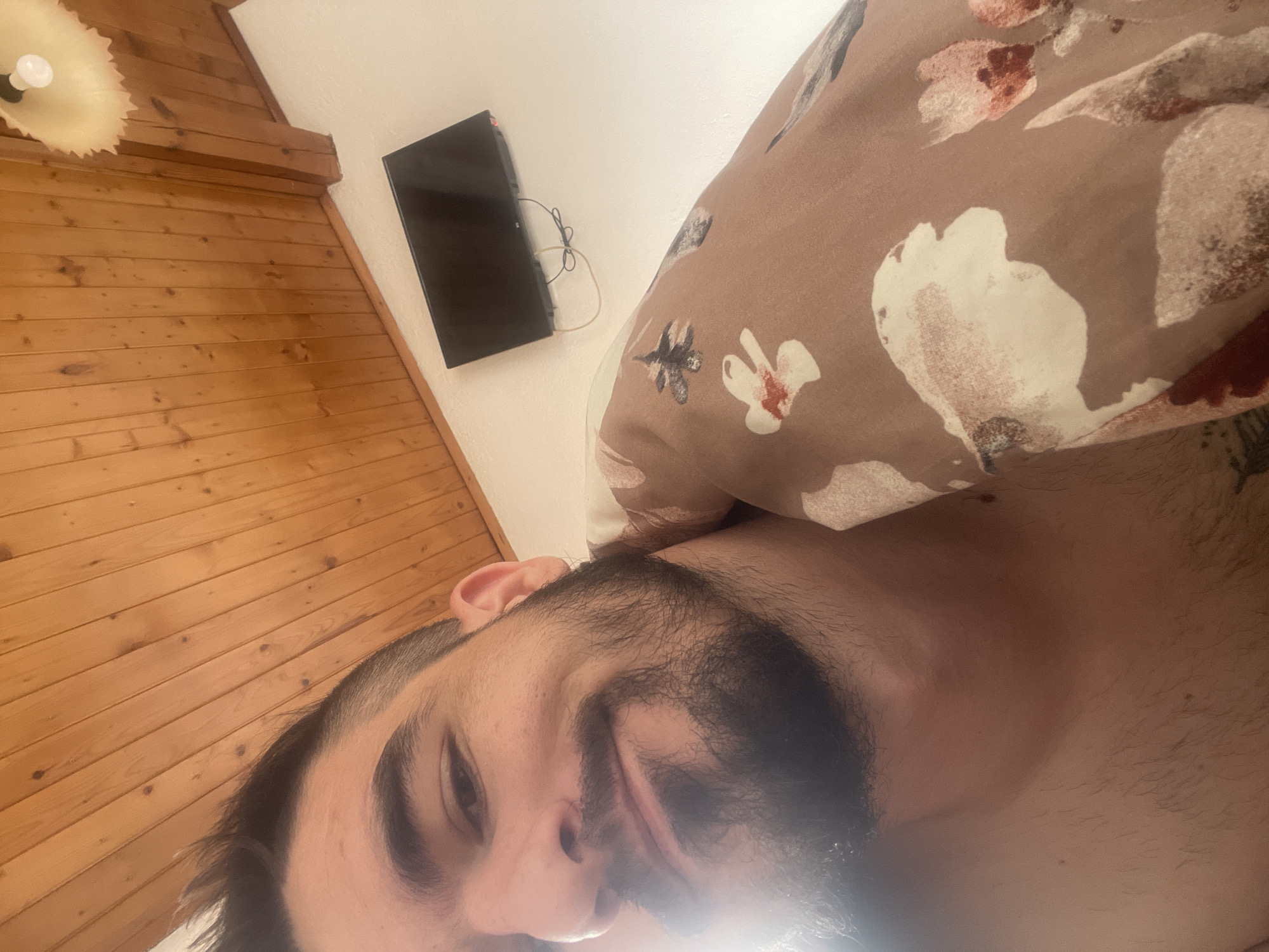 A low-angle, close-up, indoor selfie shows a person lying in bed. The person has dark hair, styled upwards, and a beard. The person is wearing a patterned duvet cover. The background shows a wooden ceiling with visible planks, a white wall, and a small black television mounted on the wall. A light fixture is visible at the top right of the image. The lighting suggests it is daytime, possibly with sunlight entering the room. The location is Unknown, Unknown.